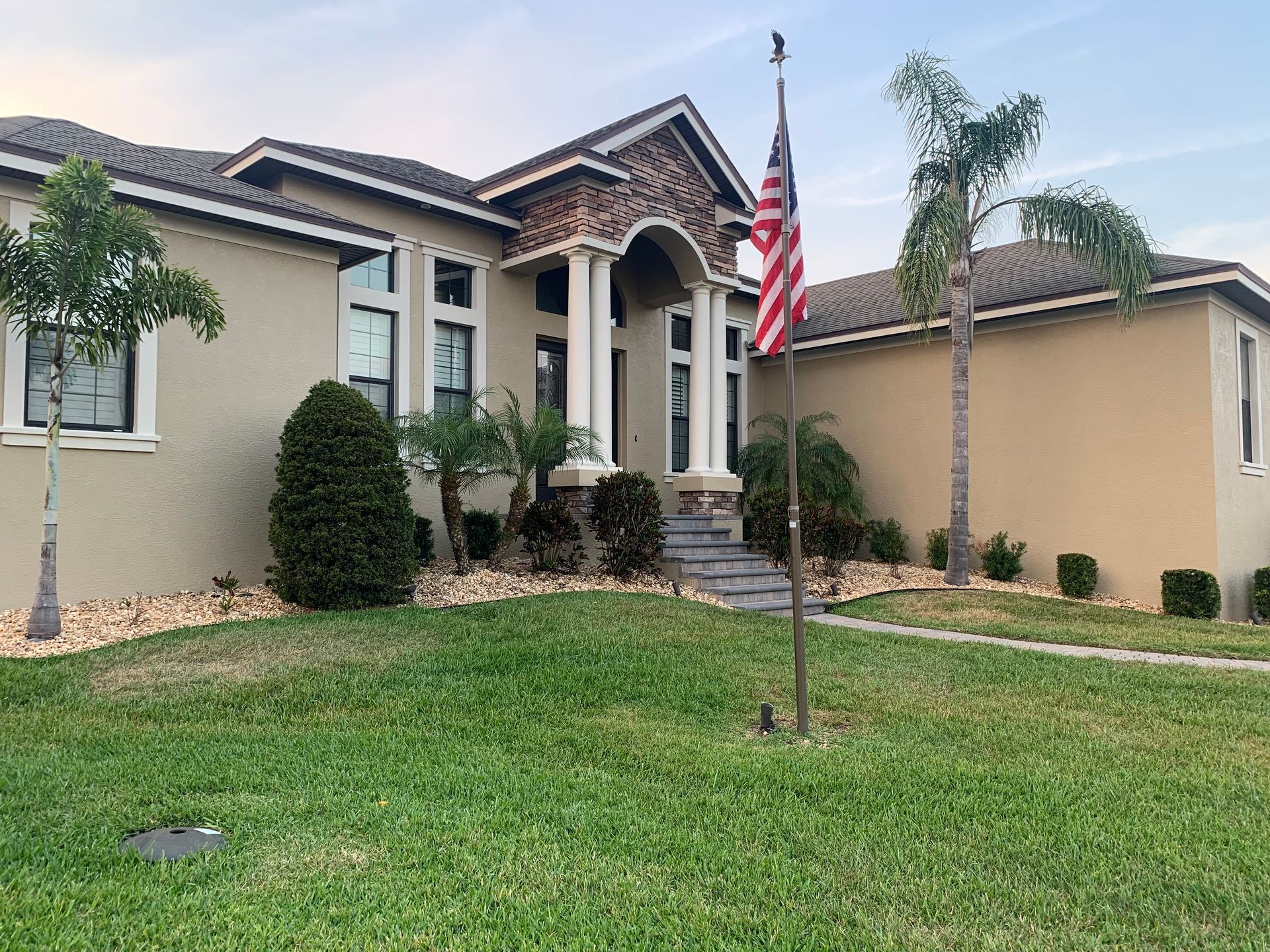 A beige house with an American flag, pillars, and palm trees in the yard.