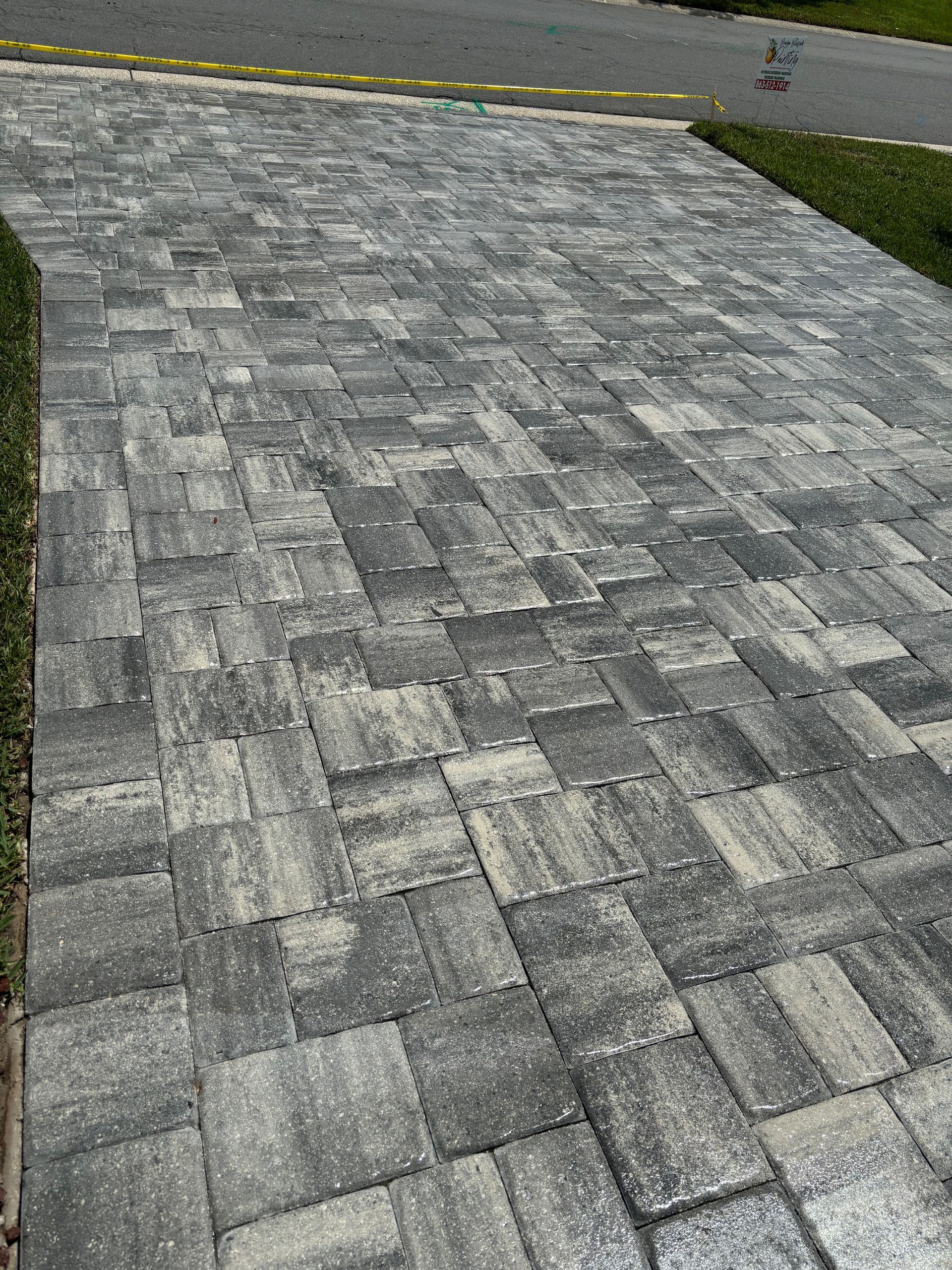 Driveway paved with gray and tan rectangular bricks, with grass on the left and a street in the background.