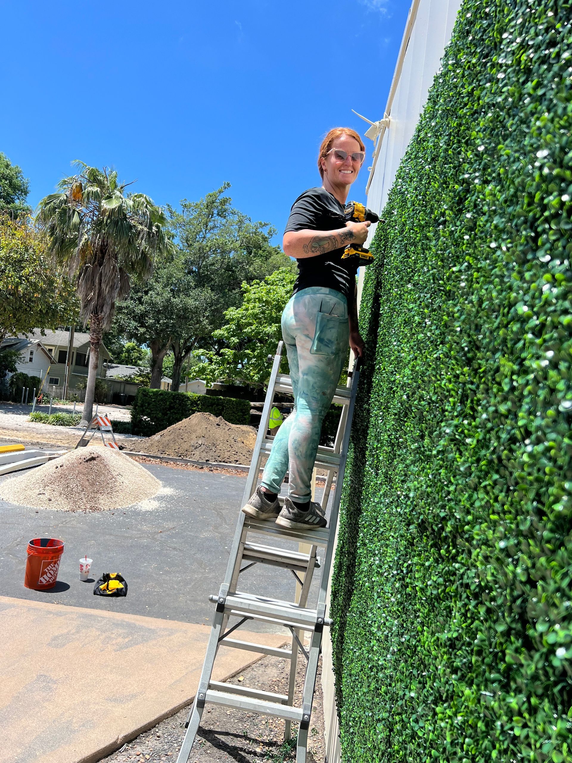 Woman on a ladder installing green wall panels, holding a power drill on a sunny day.