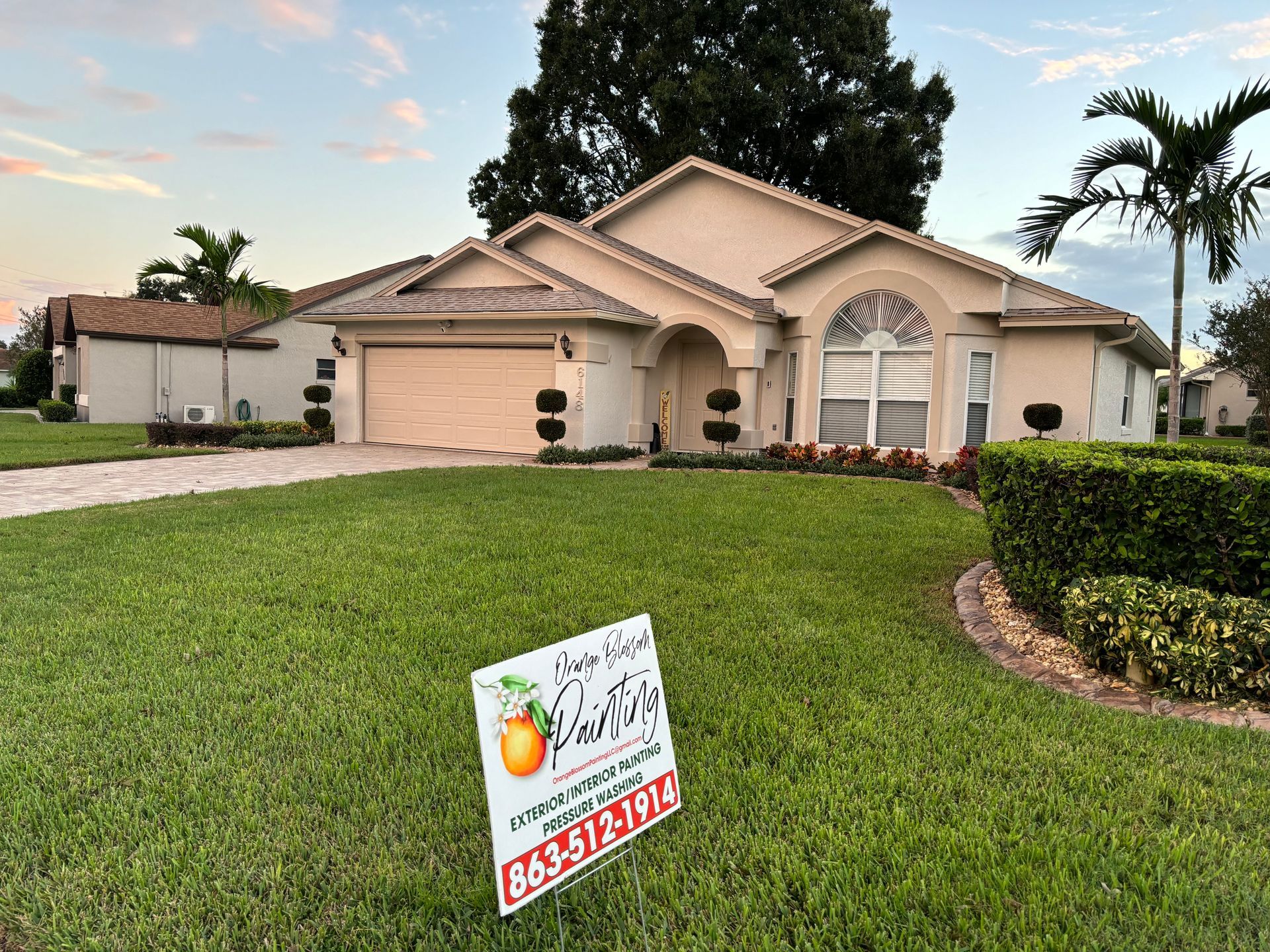 House with sign in yard, grass lawn, beige exterior, arched entryway, and palm tree.