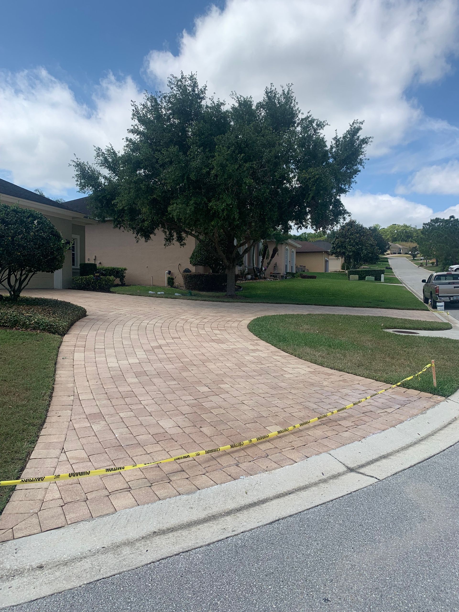 Brick driveway curves past lawn, tree in front of a light-colored house on a sunny day with blue sky.