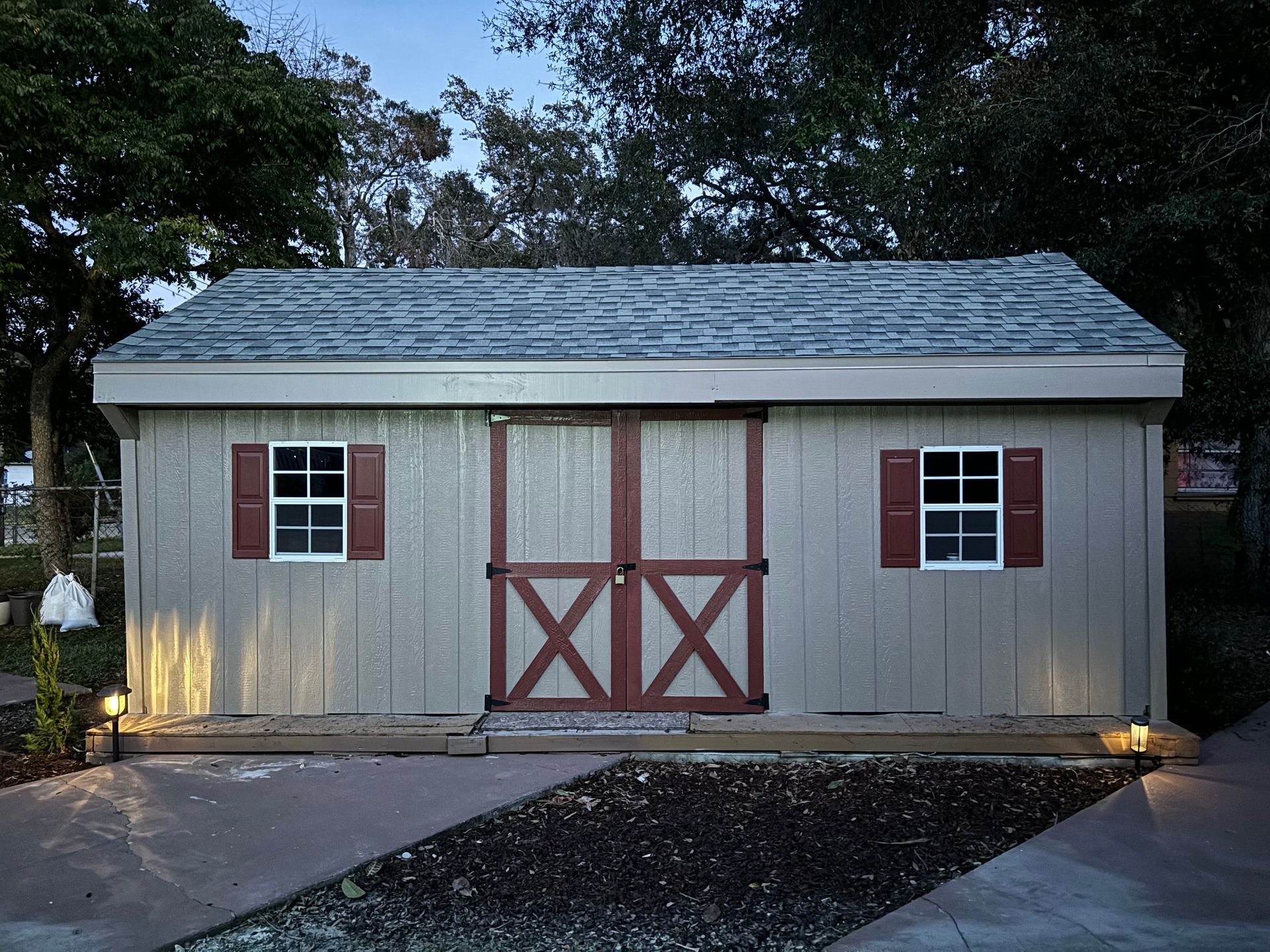 Tan shed with a gray roof, red doors, and shutters, illuminated by ground lights.