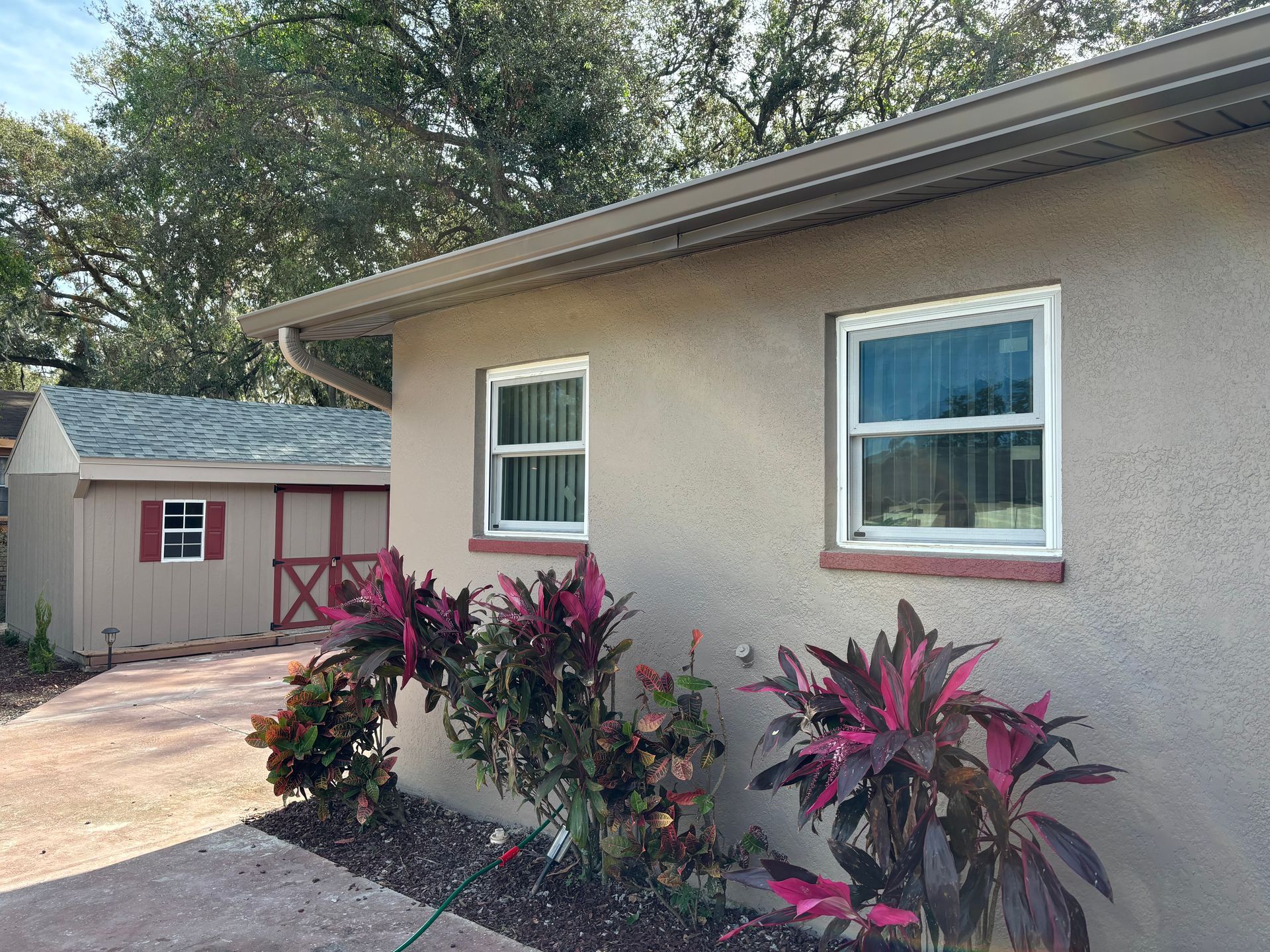 Beige stucco building with two windows, red plants, and a shed.