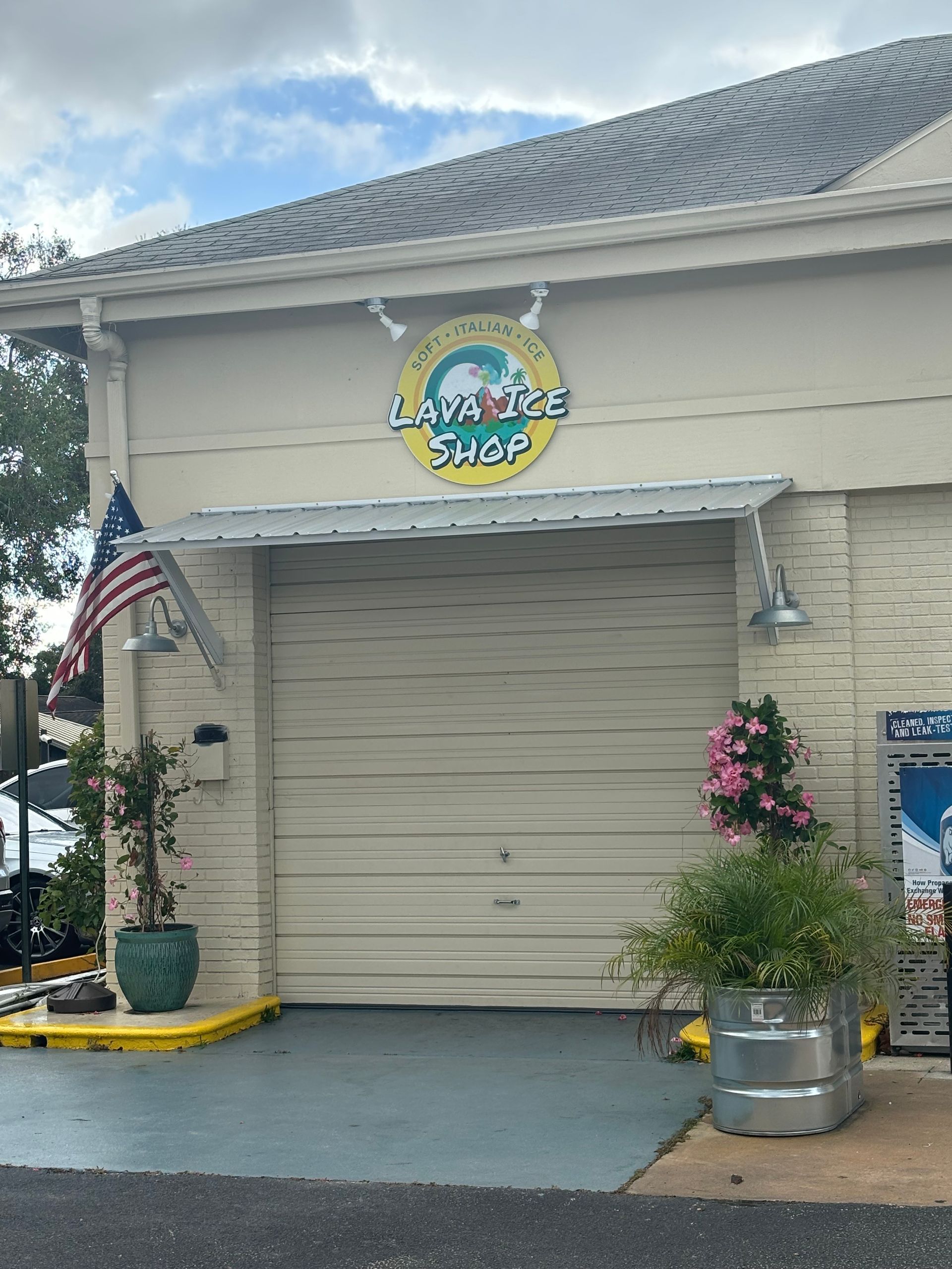Laundromat shop with a closed garage door and American flag, plants in pots, and a round sign above.