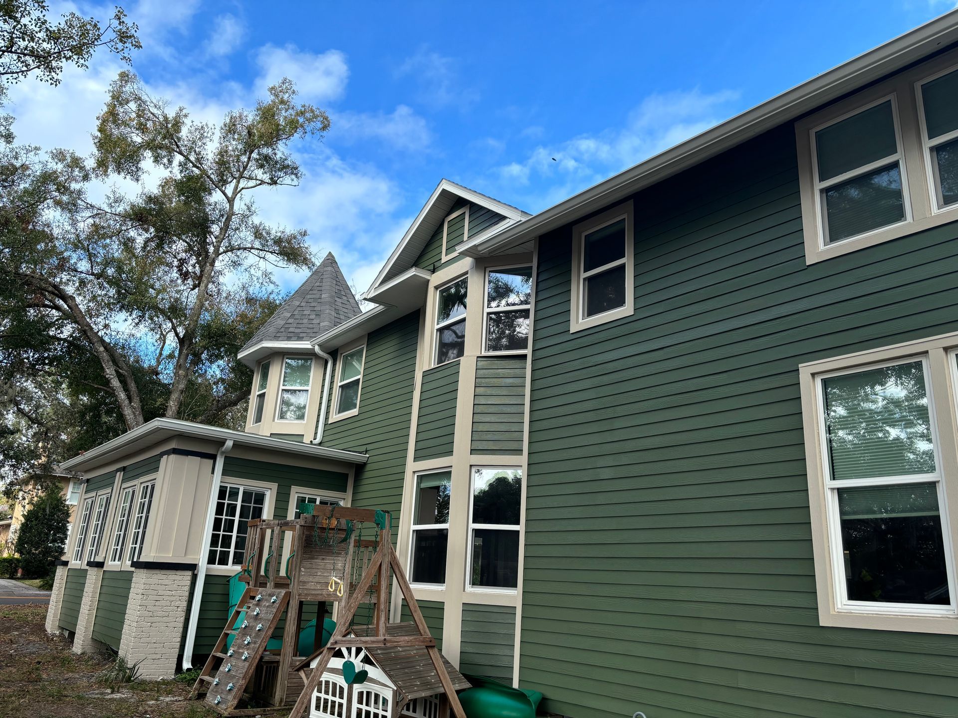 Green house with multiple windows, a turret, and a wooden play structure. Cloudy sky.
