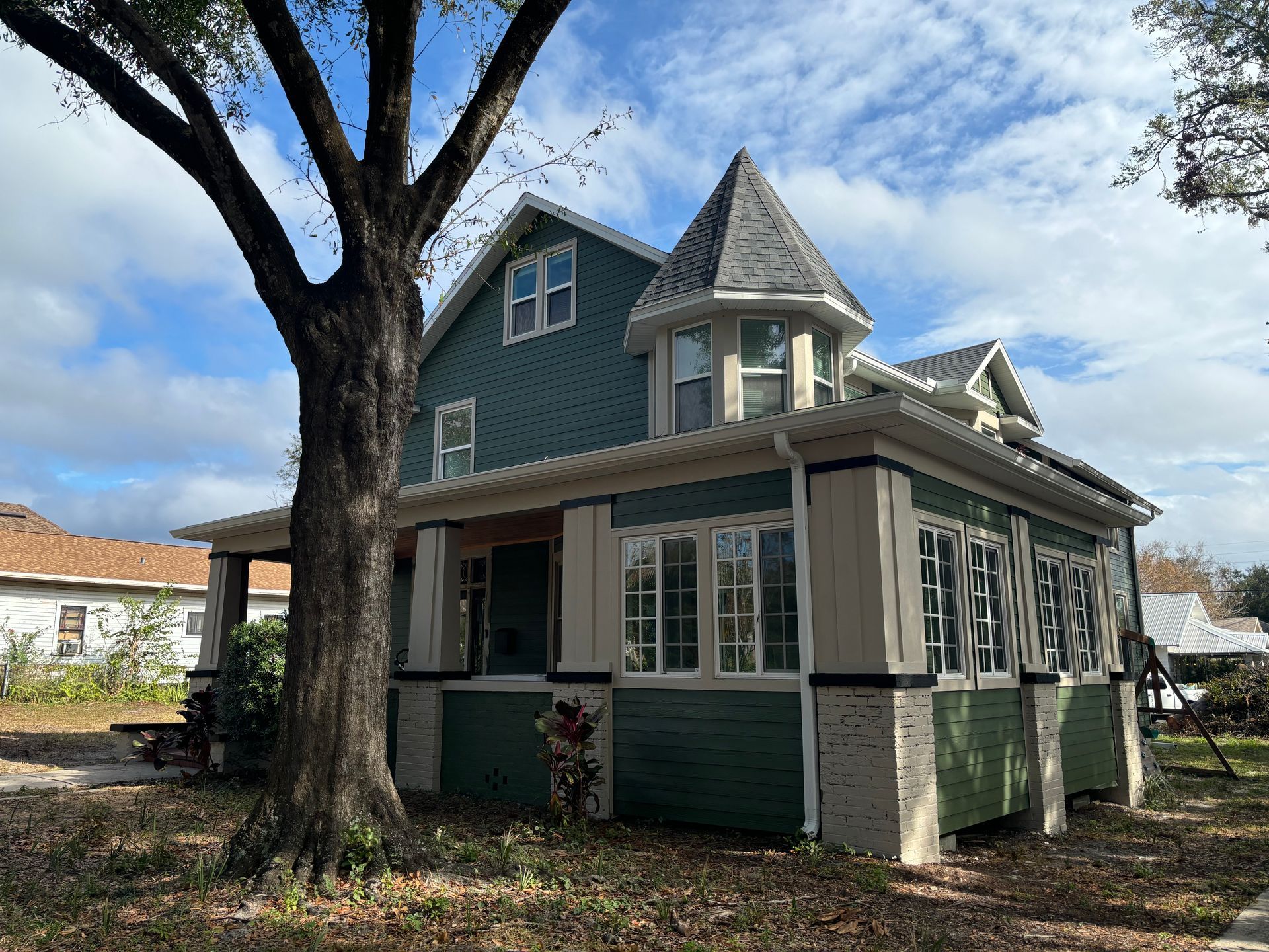 Green Victorian house with turret, white trim, and porch, under a blue sky with a large tree in front.