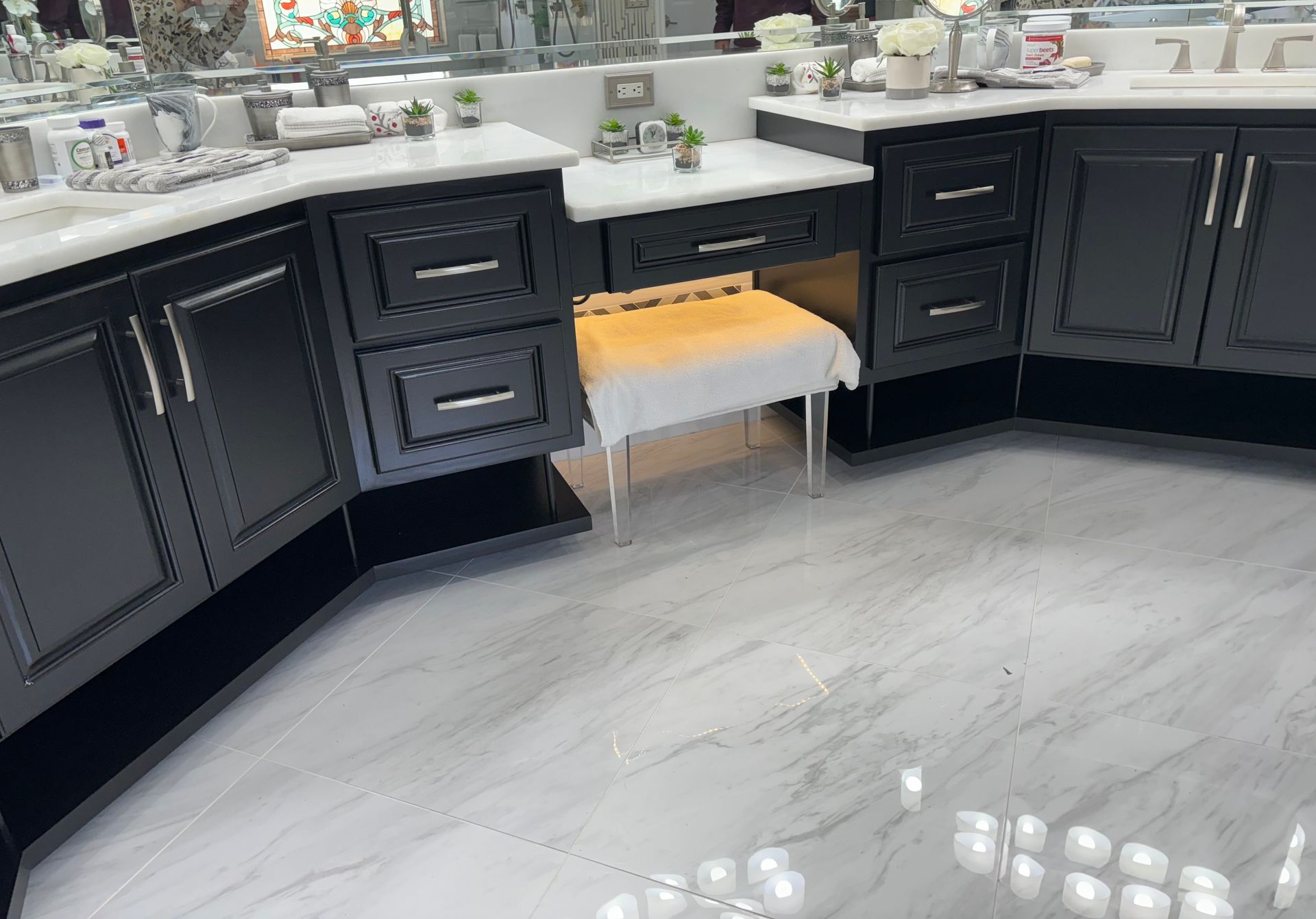 A bathroom with black cabinets , white counter tops , and a stool.