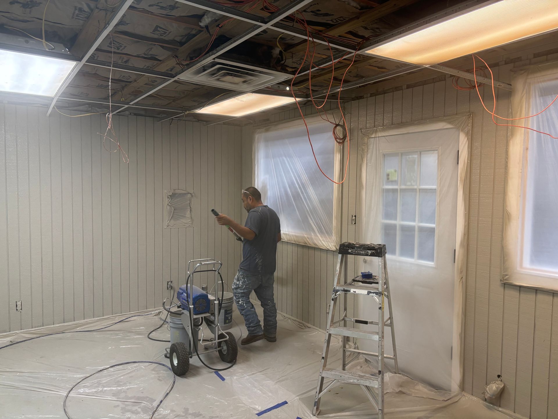 Man painting a room with a paint sprayer. Walls are light beige. Door and windows are covered.