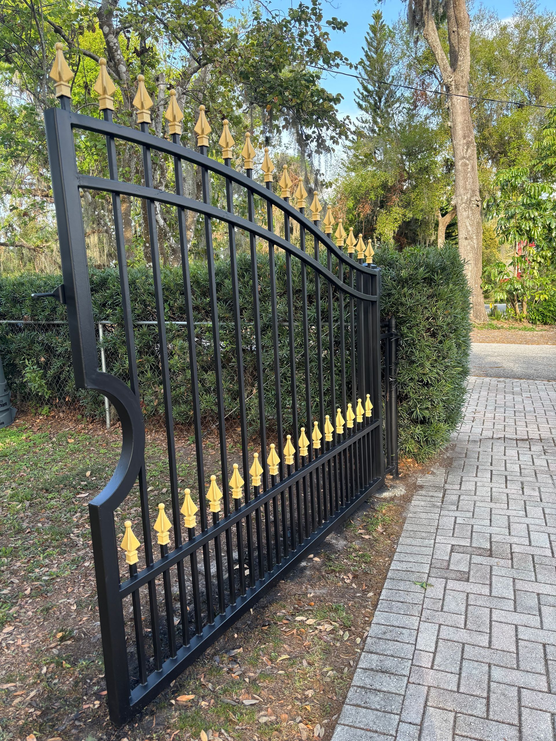 Black and gold wrought iron gate ajar, set on a brick pathway, with green bushes and trees in the background.