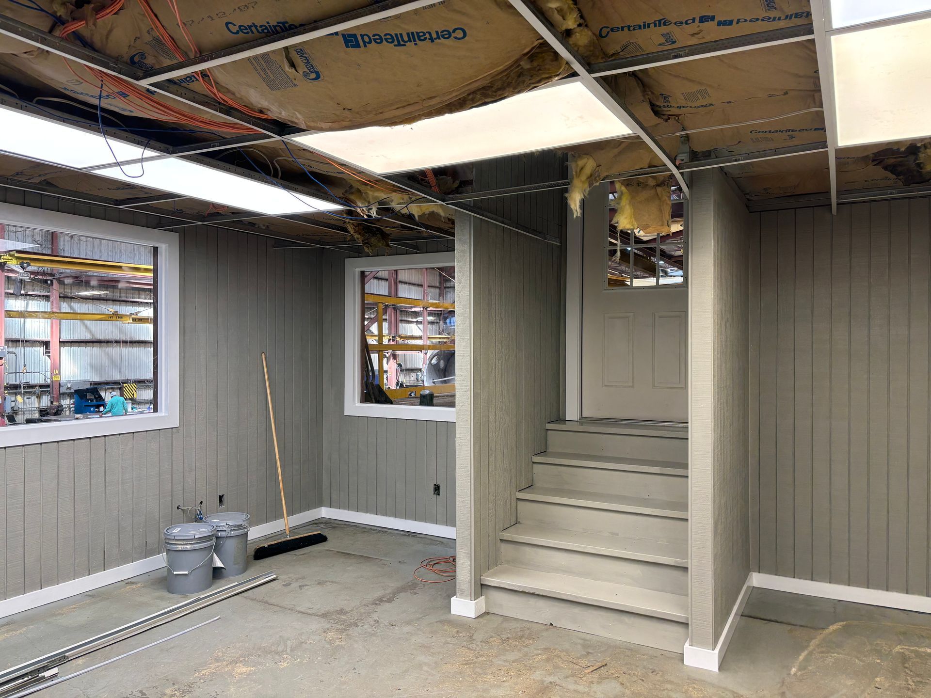 Interior of a room under construction, with gray paneling, stairs, windows, and a door.