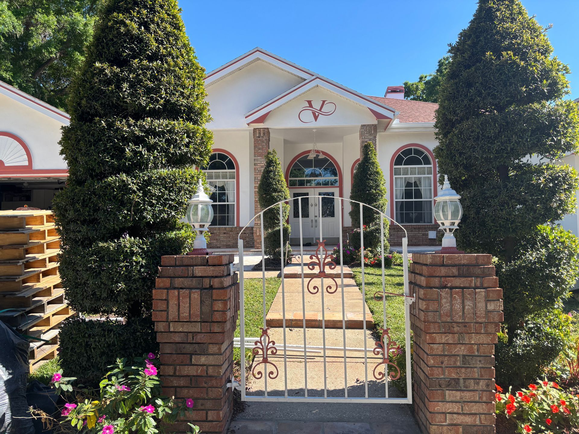 White house with arched windows, brick pillars, and iron gate. Two tall, manicured bushes frame the entrance.