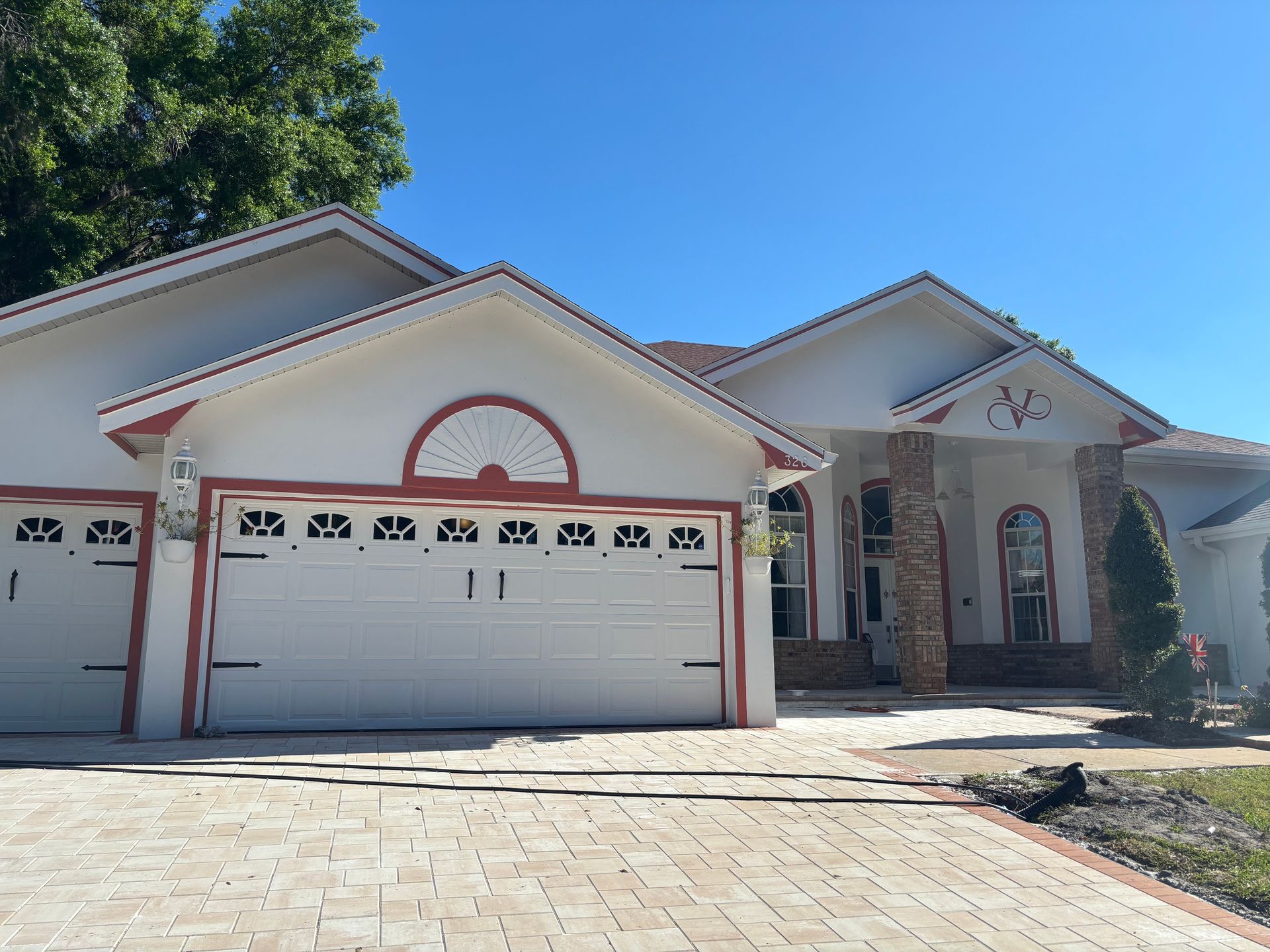 White stucco house with red trim, two-car garage, arched windows, and brick driveway on a sunny day.