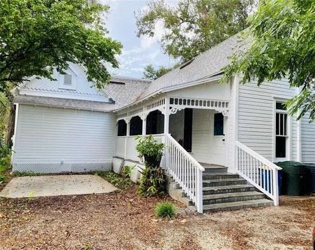 White house with porch, steps, and greenery in a yard. Cloudy sky above.