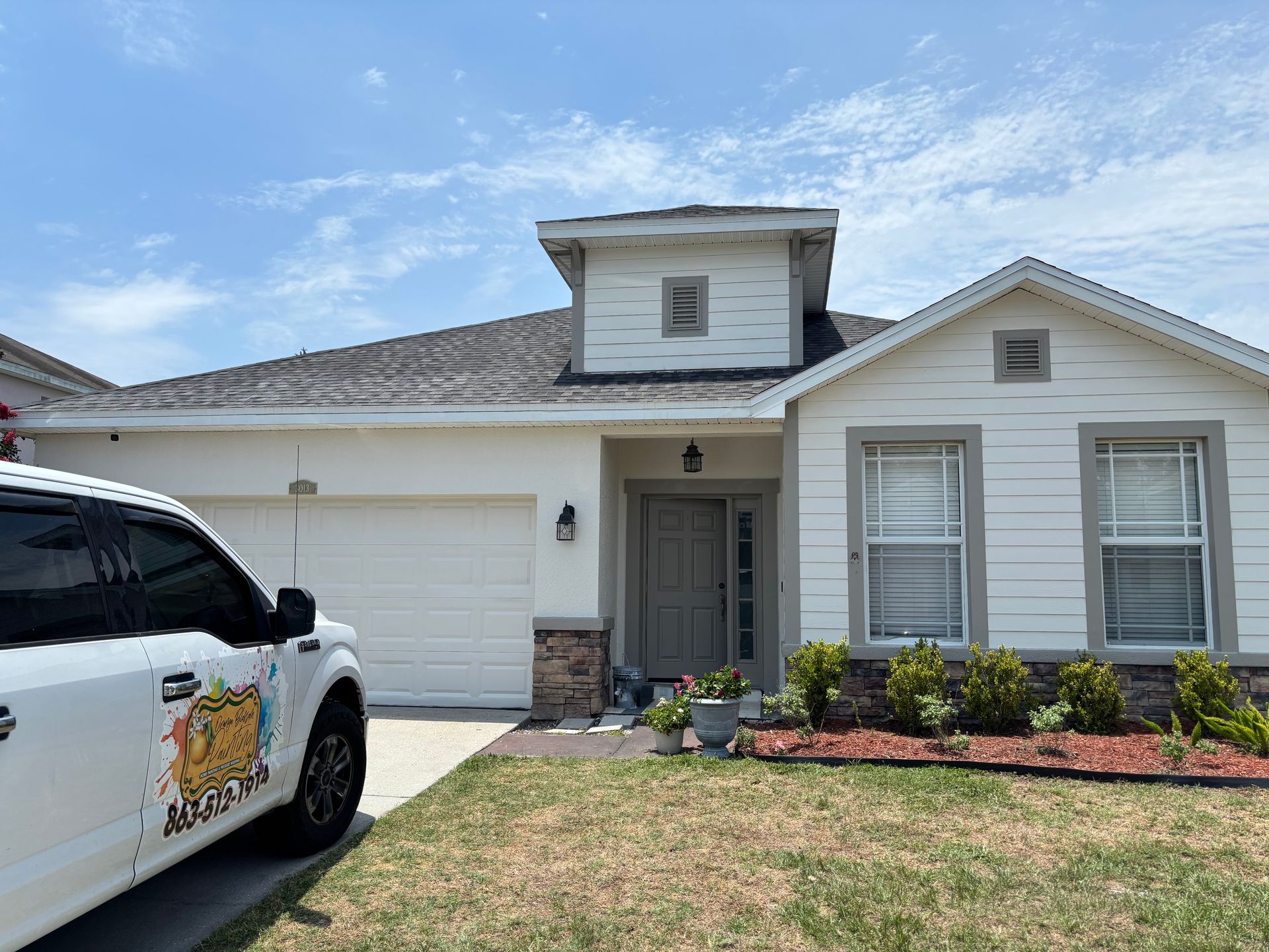 White house with a truck parked in front. Blue sky, plants, and a garage door.