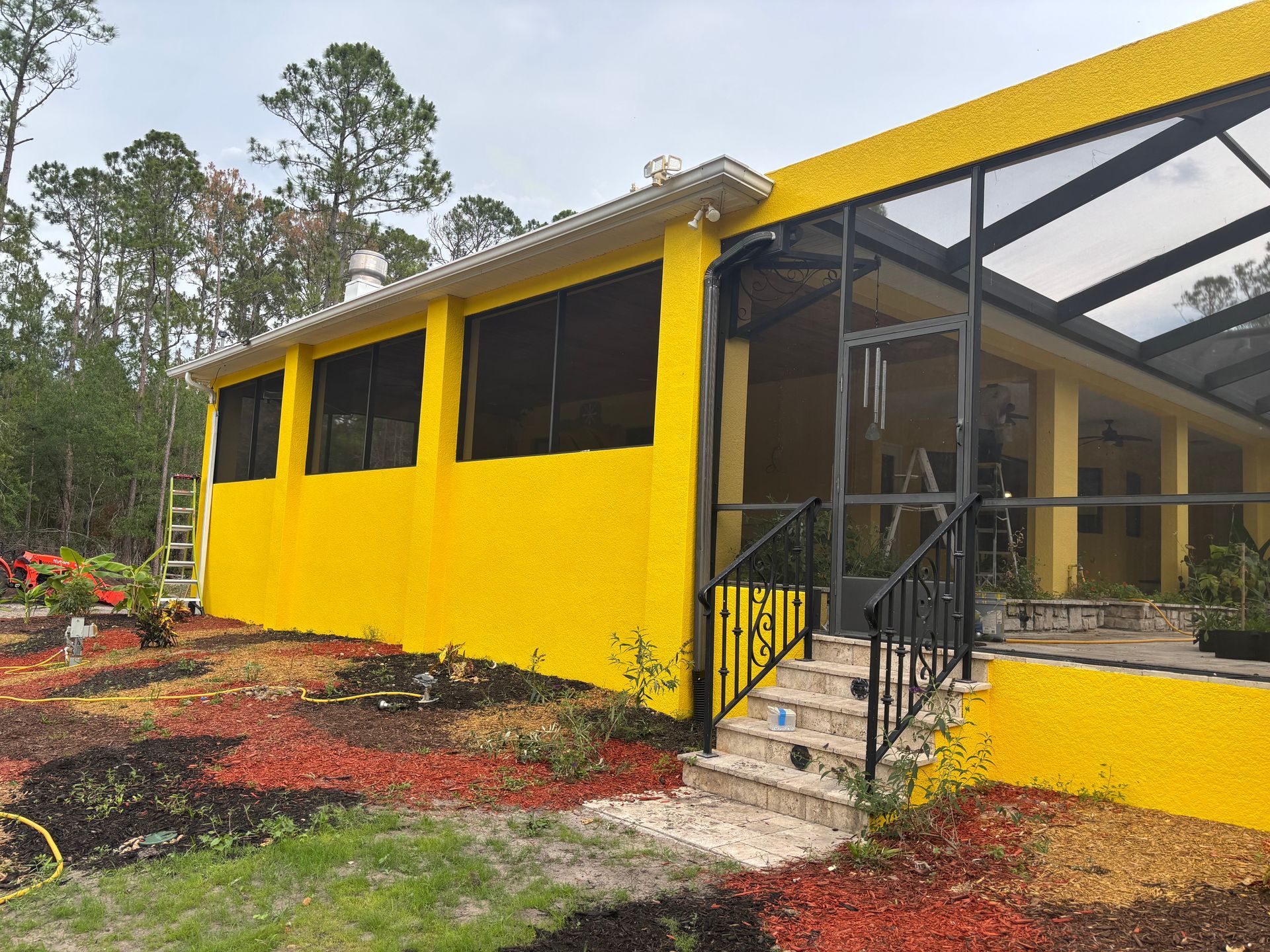 Yellow stucco house with black screened porch and steps. Landscaping with red mulch.