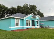 Aqua-colored house with white trim and gray roof against a backdrop of green trees under a cloudy sky.