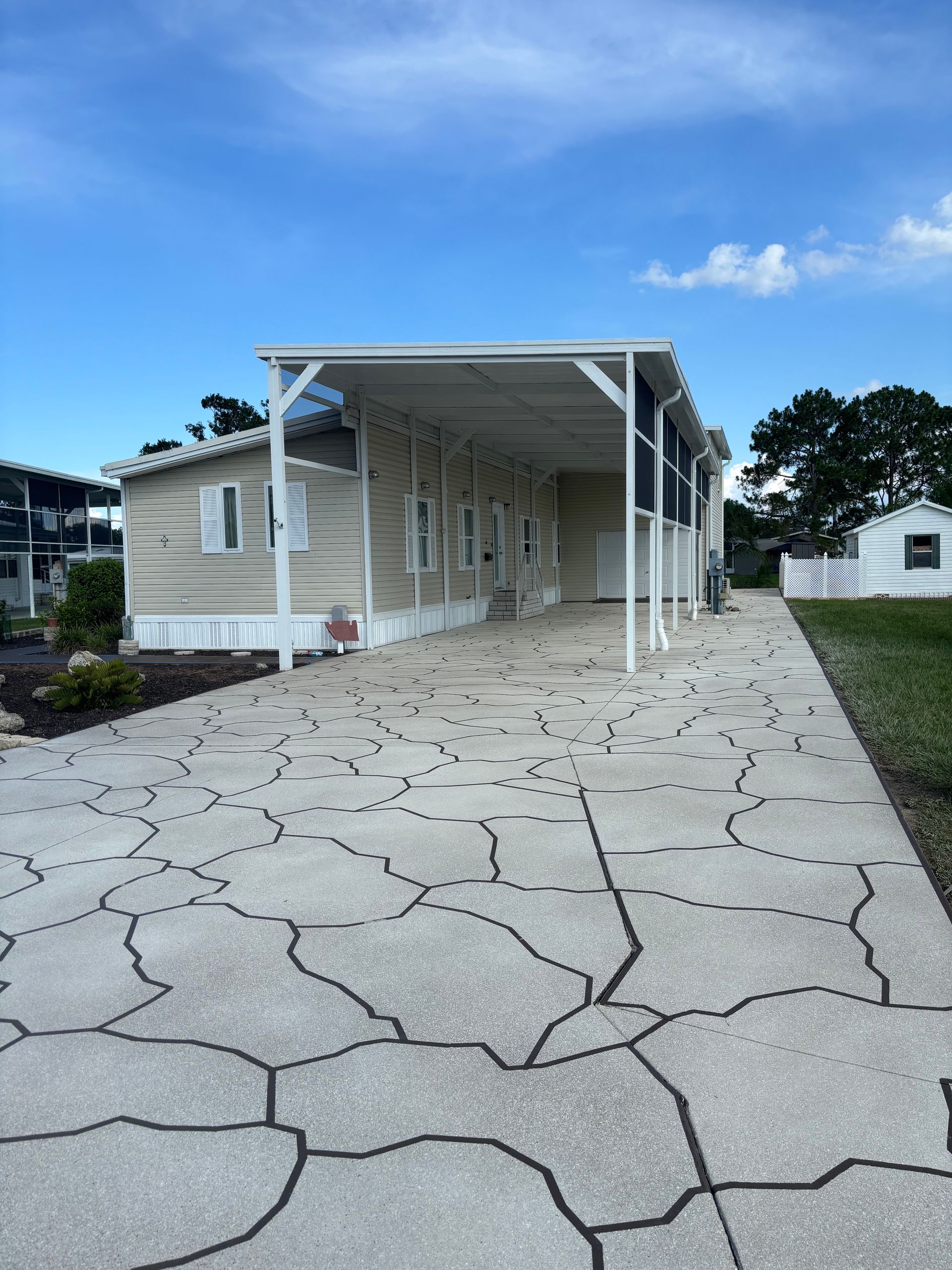Tan mobile home with a white carport and patterned driveway under a blue sky.