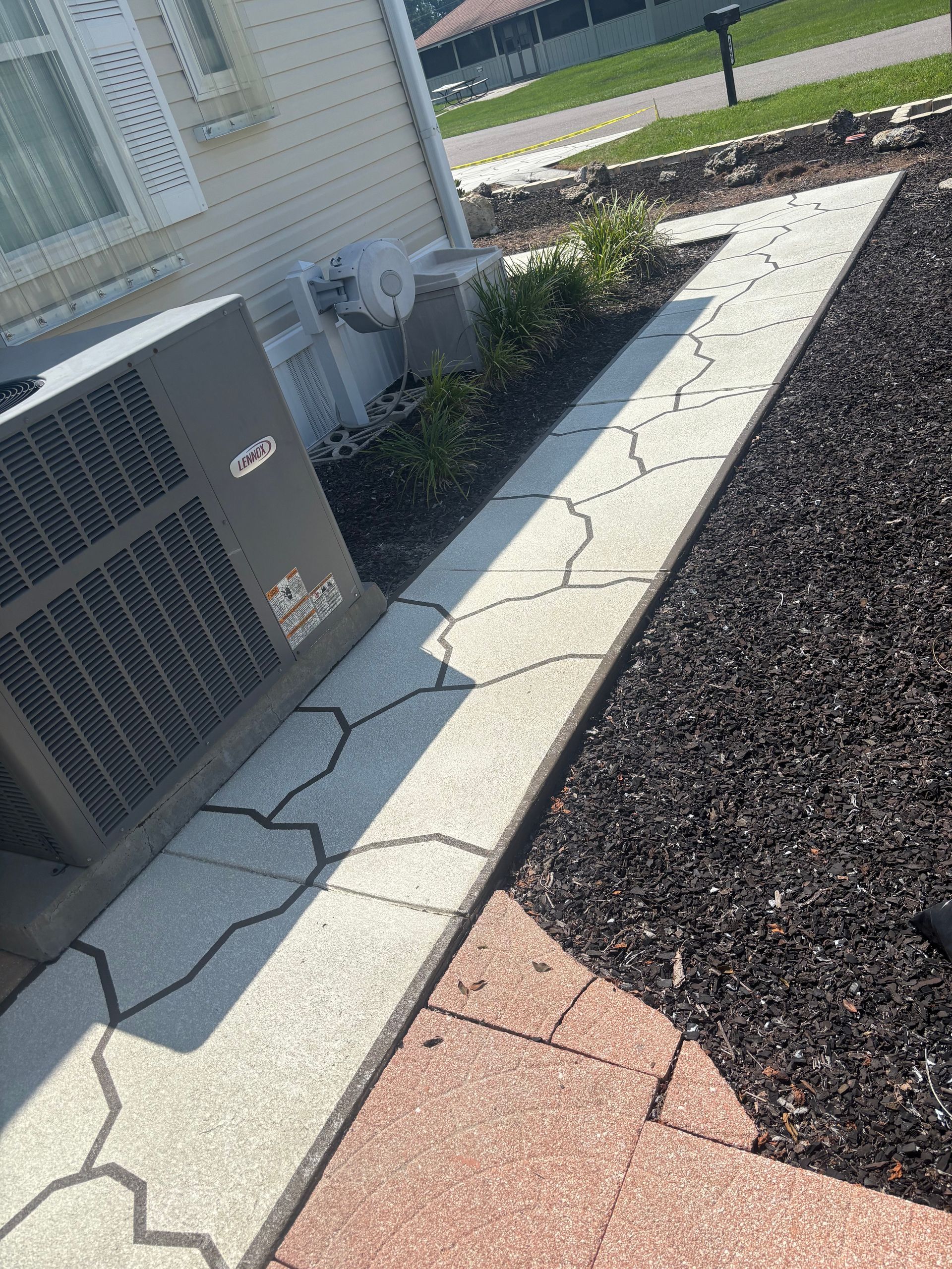 A concrete walkway with cracked, decorative edges, next to a home's air conditioning unit and dark mulch.
