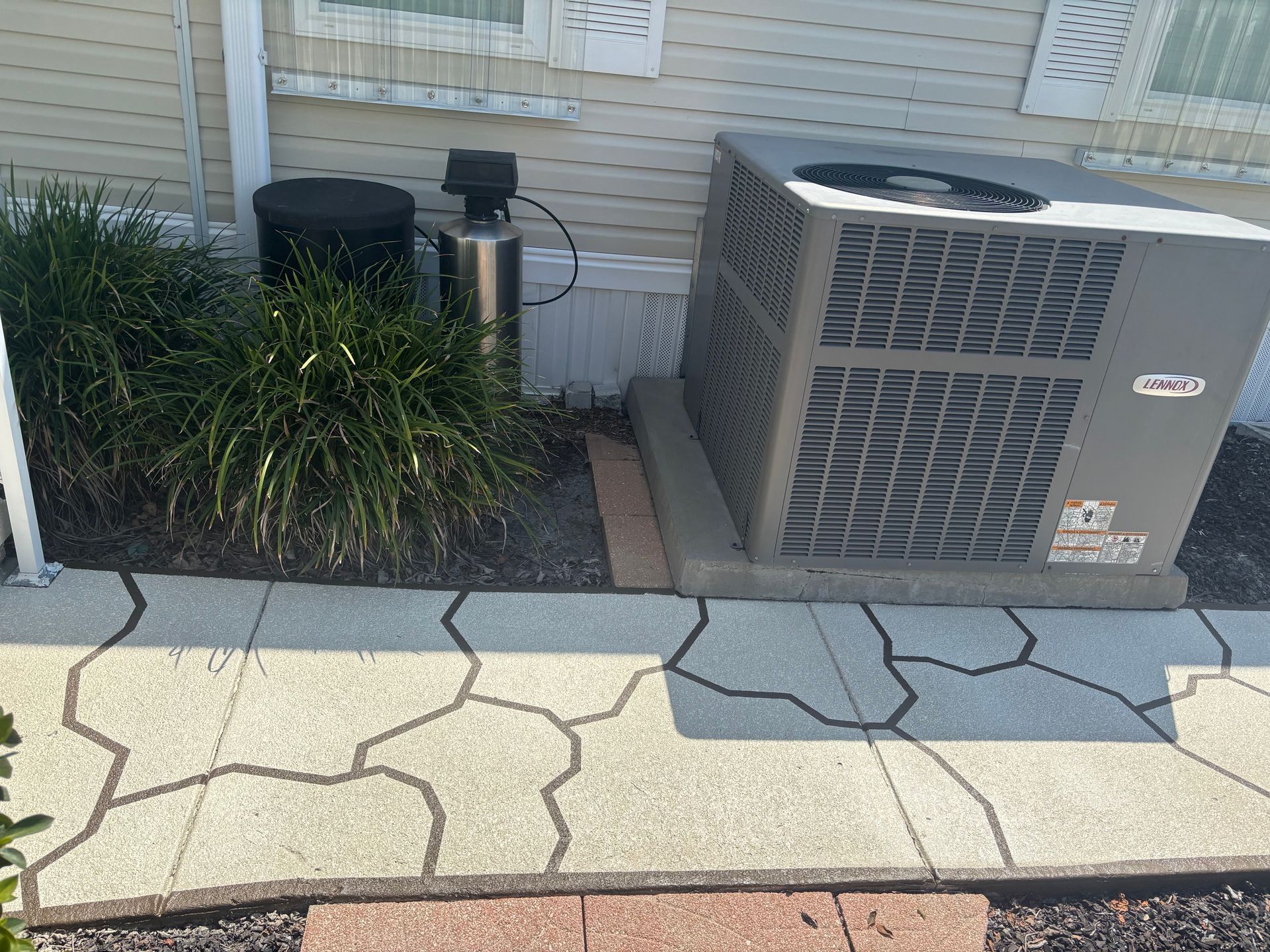 Air conditioner, water softener, and shrubbery next to a house with a patterned concrete walkway.