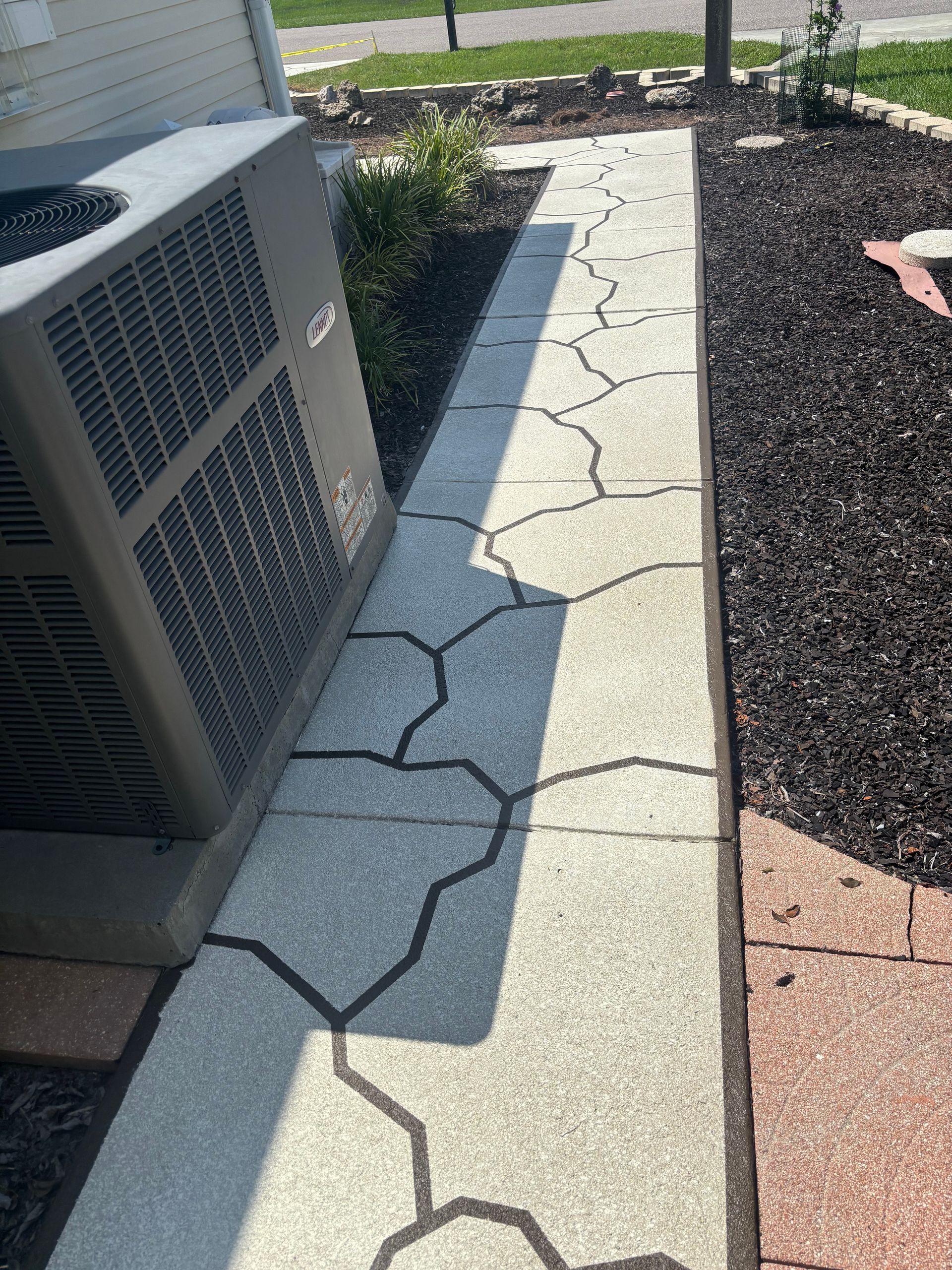 A concrete walkway with irregular black lines, beside an air conditioner unit and a bed of dark mulch.