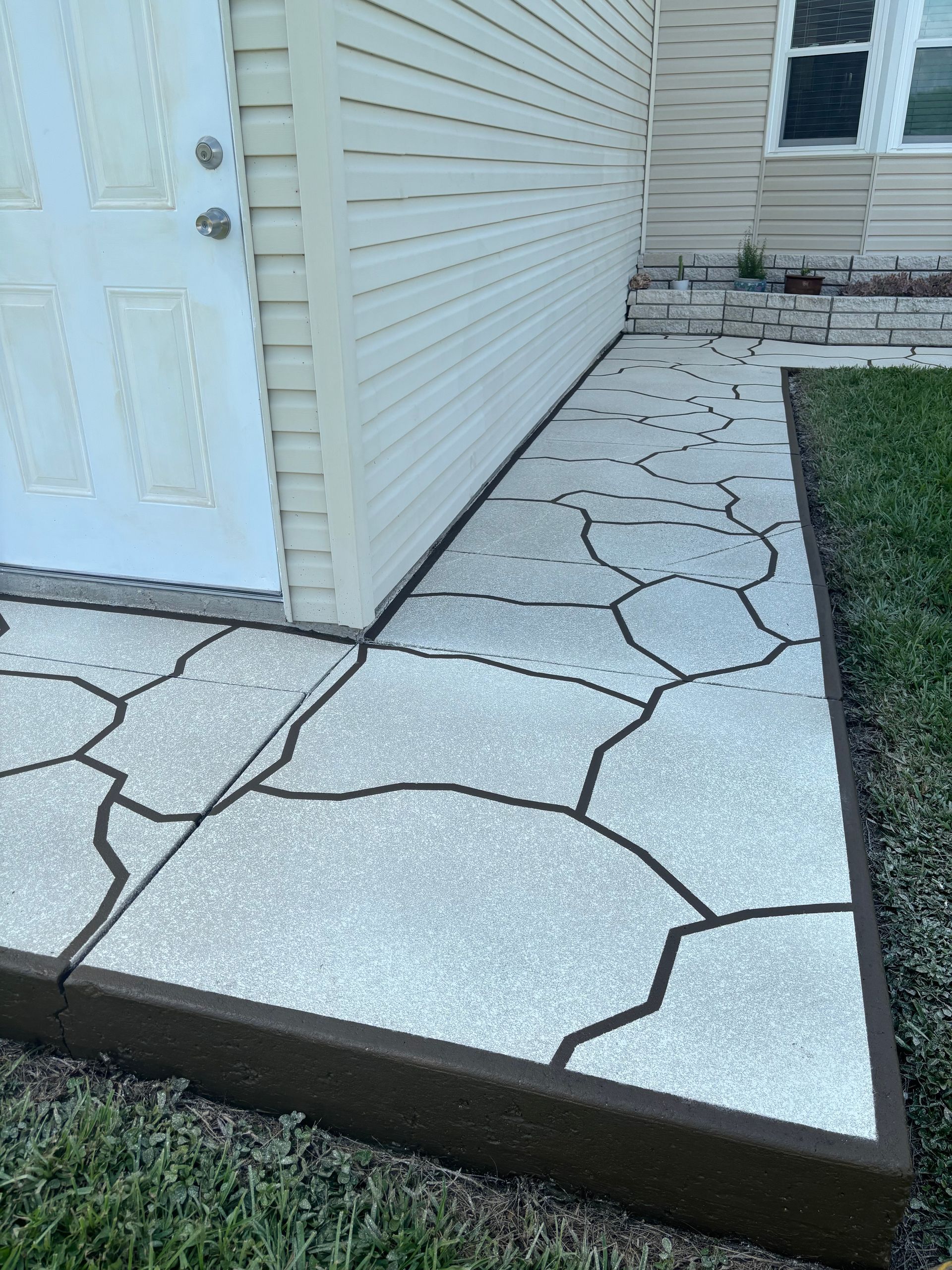 Concrete walkway with decorative stone pattern, brown border, next to building and grass.