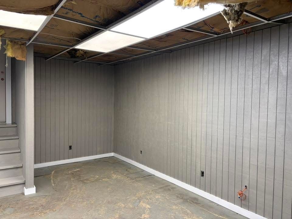 Gray-paneled room with white baseboards and fluorescent ceiling lights; unfinished concrete floor.