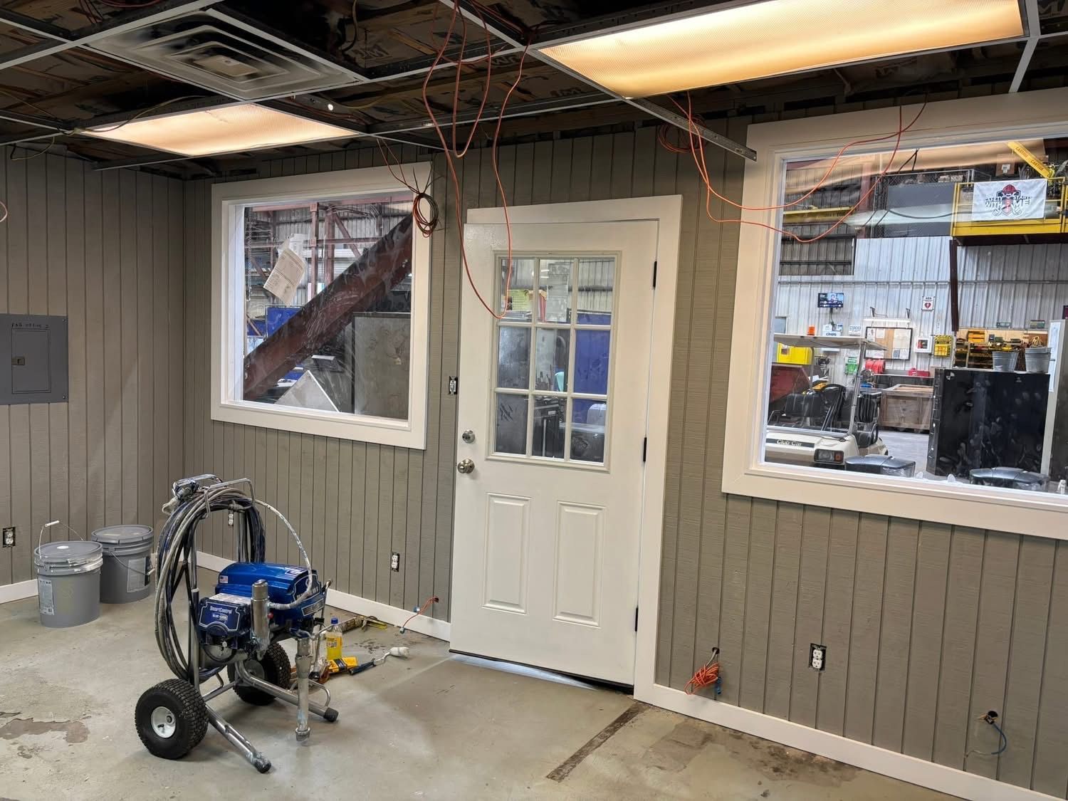 Interior room with gray paneling, white trim, door, windows; paint sprayer in foreground.
