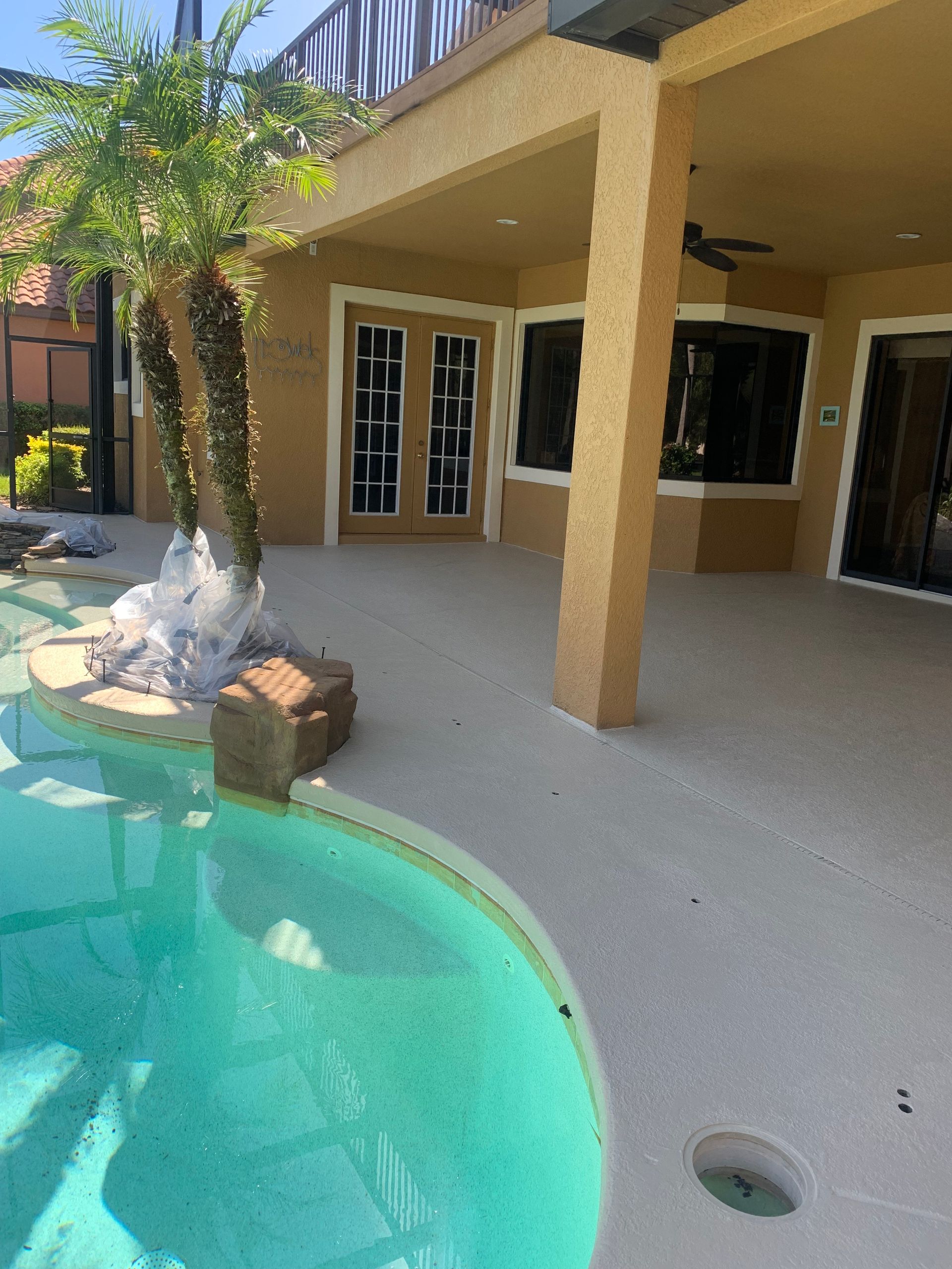 Poolside patio with turquoise water, tan deck, and light brown building. Palm tree wrapped for protection.