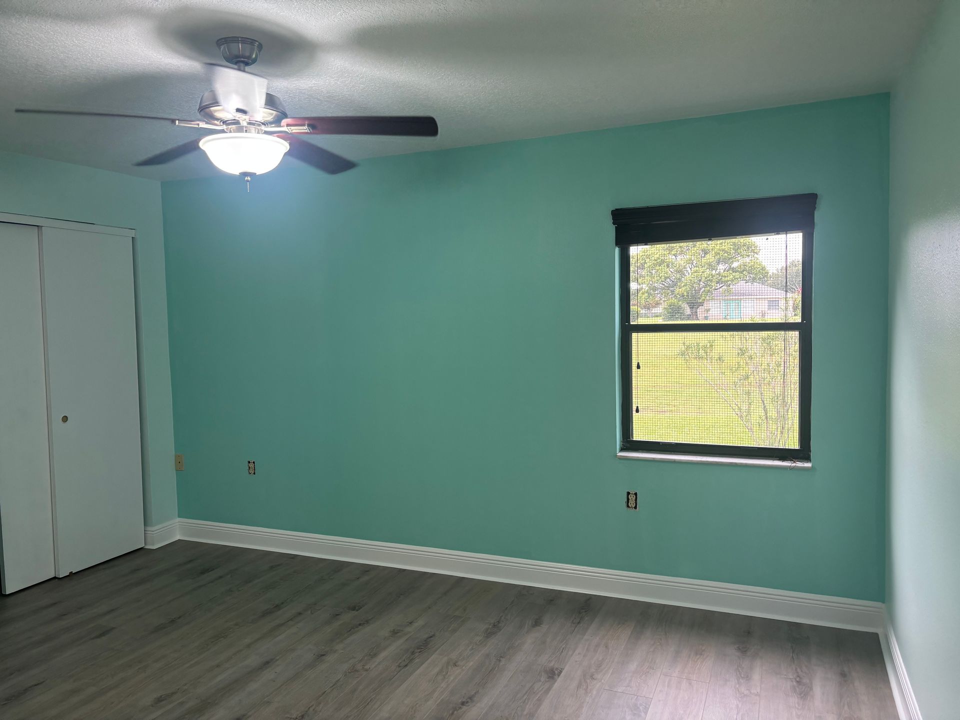 Empty room with teal walls, wood-look flooring, a window with a dark shade, and a ceiling fan.