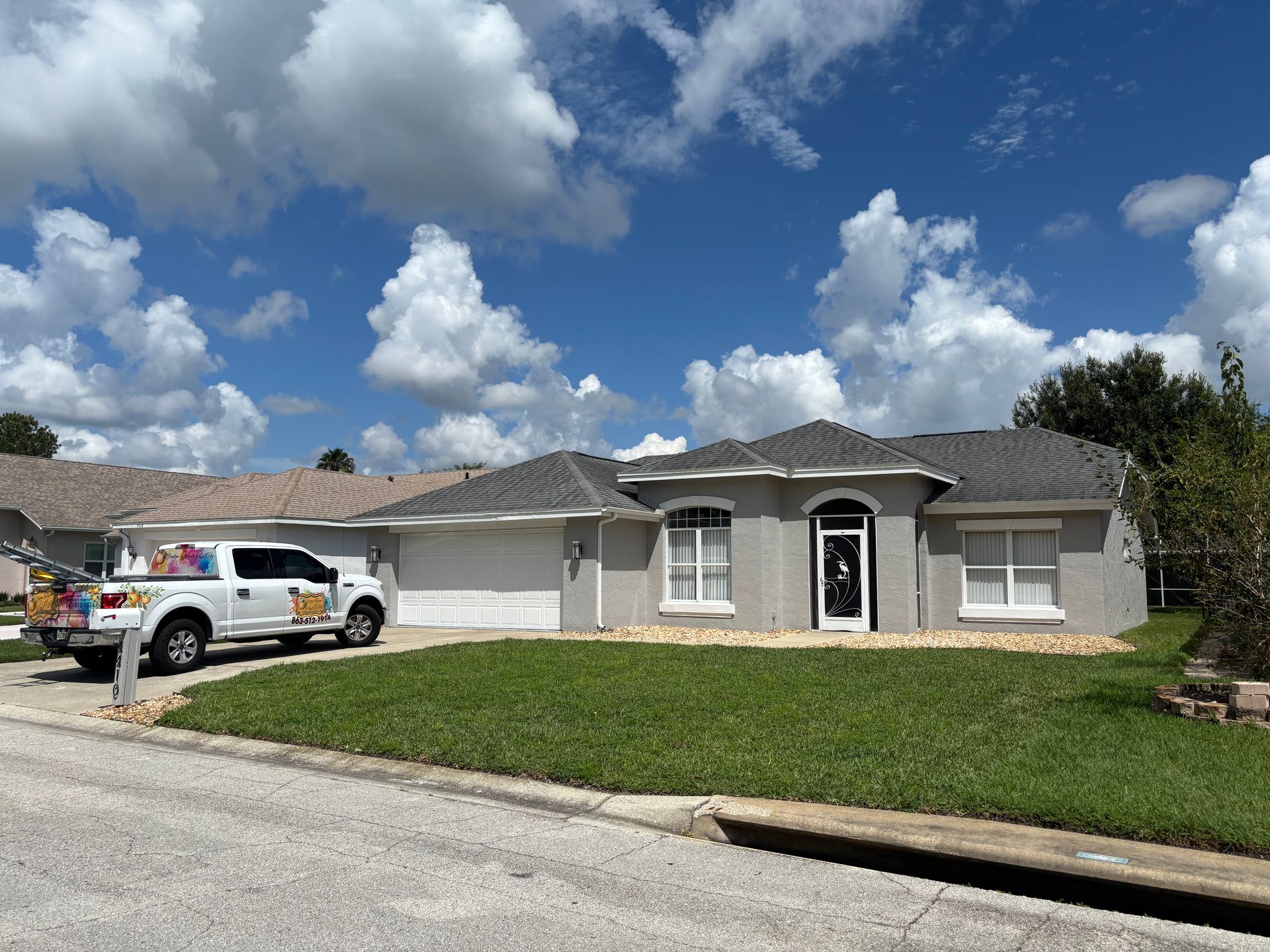 Gray stucco house with a green lawn and white truck parked in front, under a cloudy blue sky.