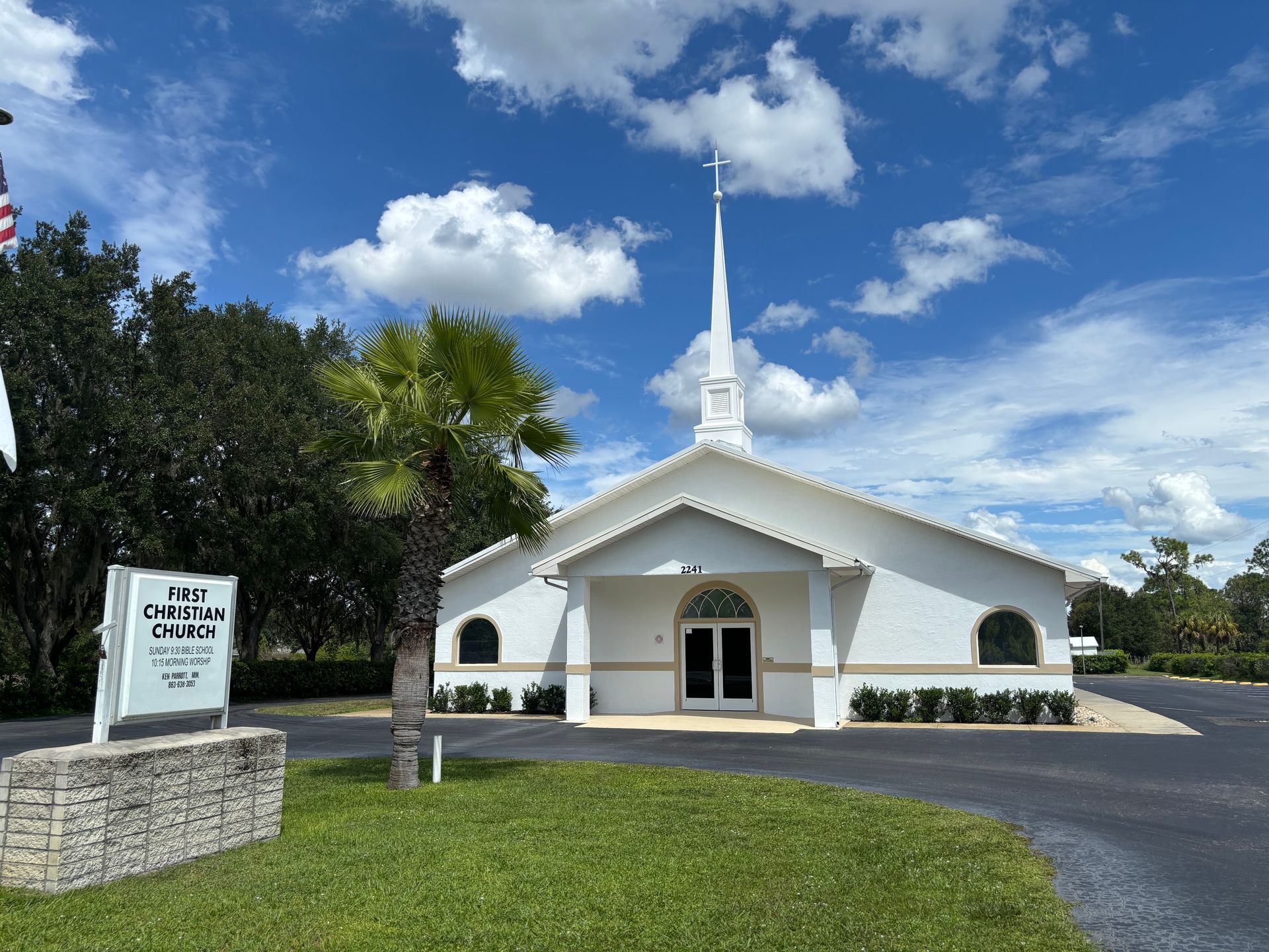 White church building under a blue sky with fluffy clouds, a sign, and green lawn.