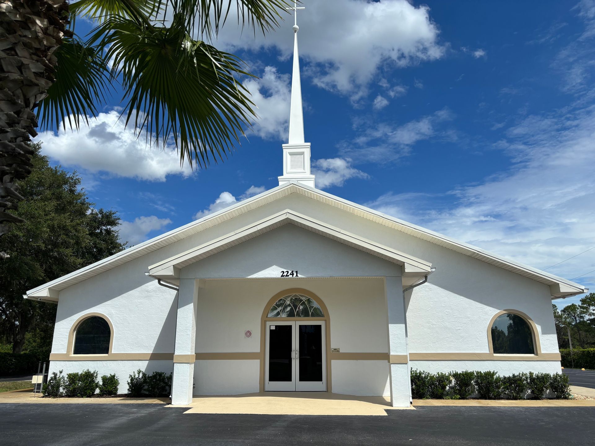 White church building with steeple under a blue sky with some palm leaves in the corner.