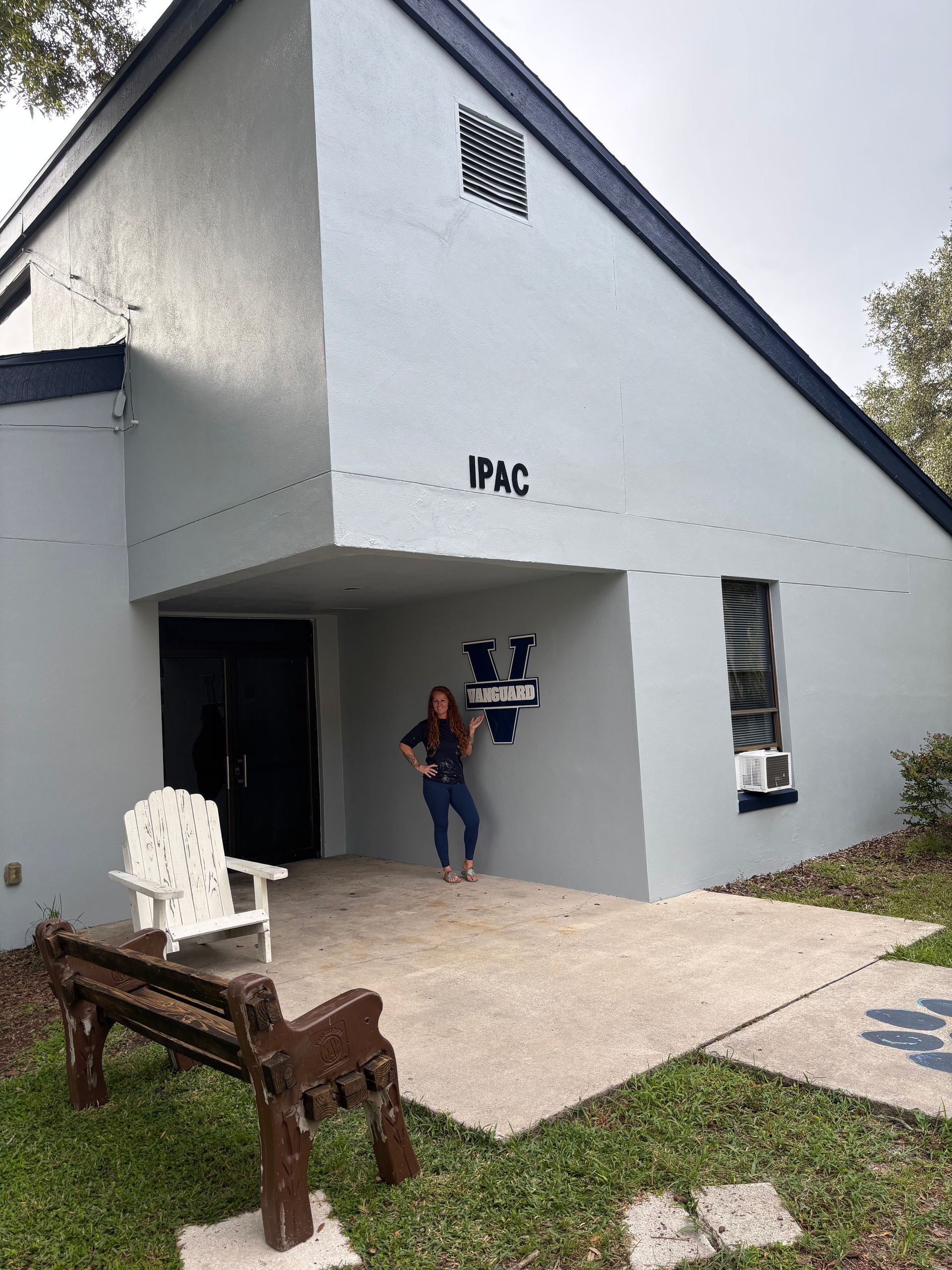 Woman standing outside a light blue building with a dark blue trim. The building has a covered entrance, bench, and chair.