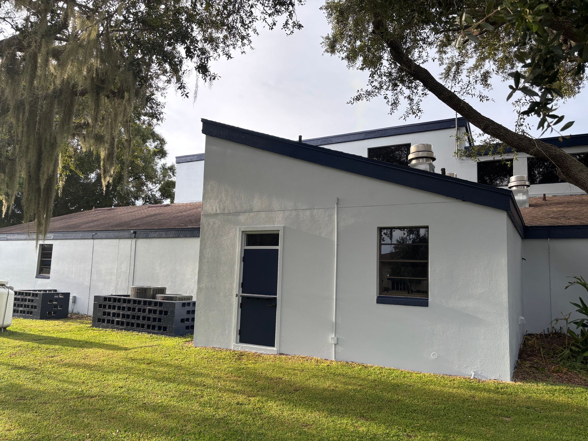 White building with blue trim, door, and window in a grassy area under a tree.
