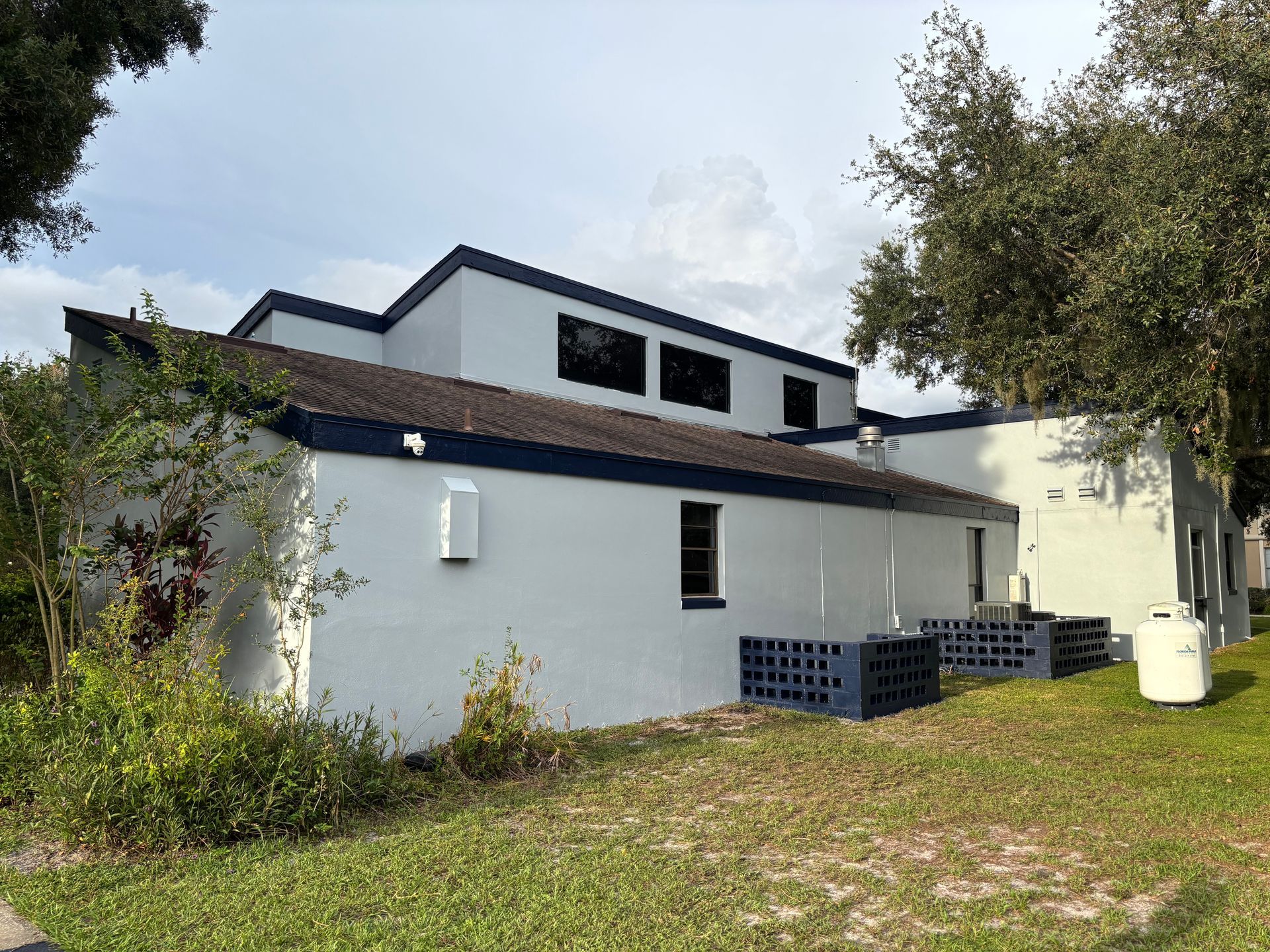 White building with dark blue trim, windows, and air conditioning units, set in a grassy area with trees.