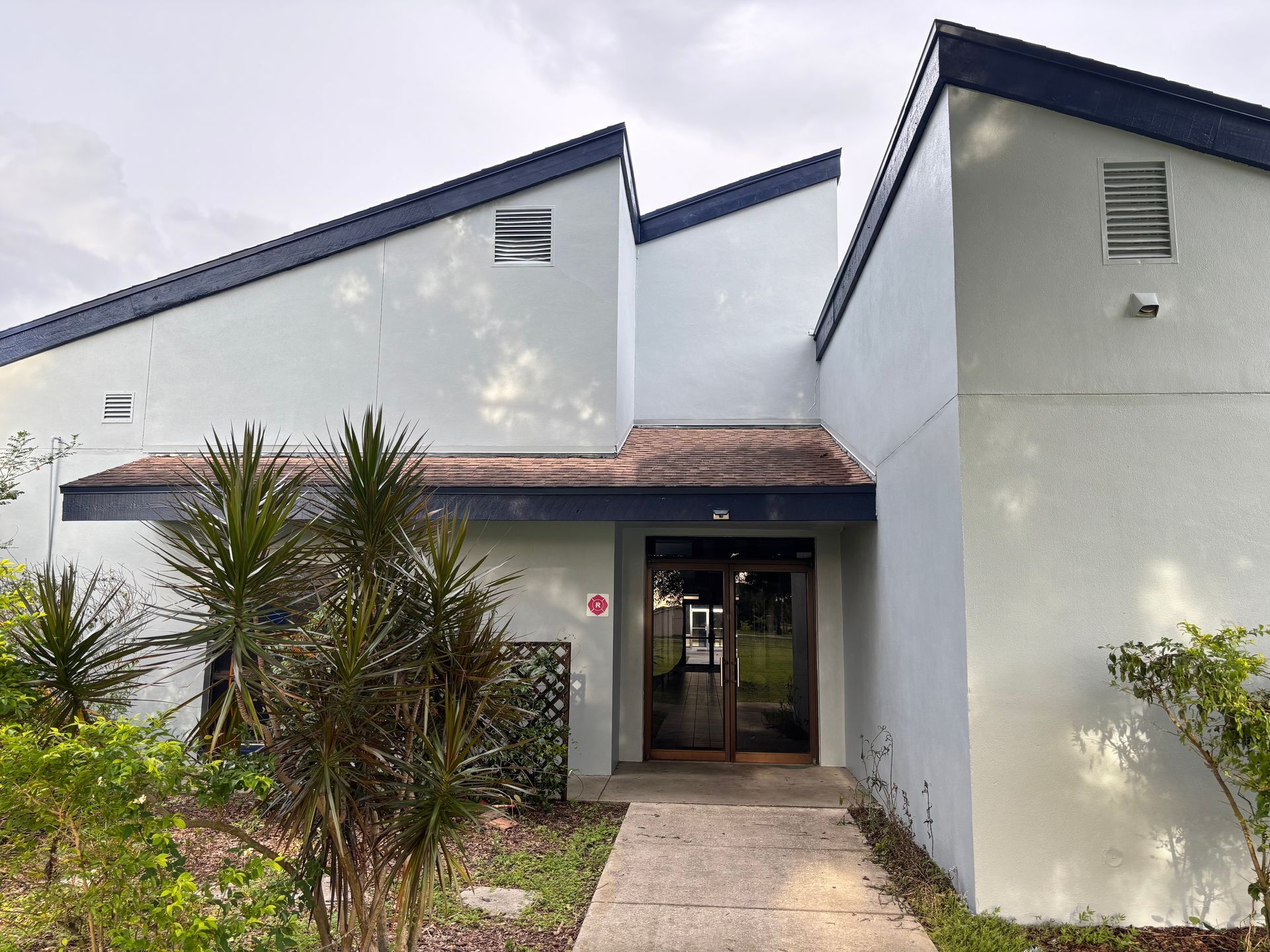 Light blue building with dark blue trim, glass door entrance, walkway, and greenery.