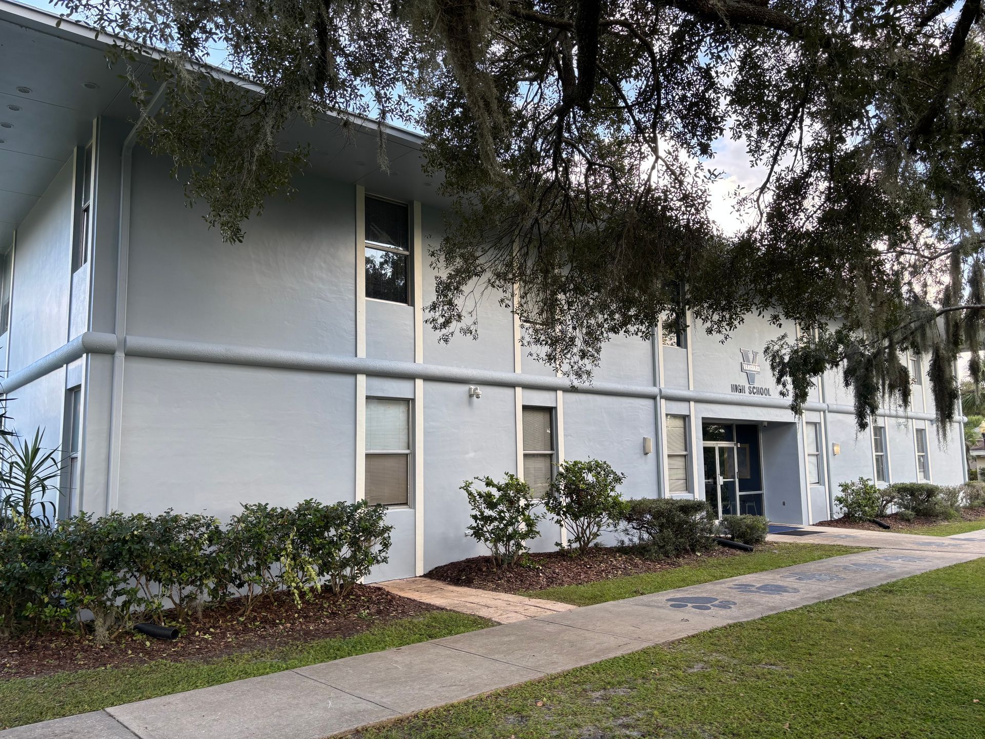 Two-story pale blue building with windows and a central entrance, surrounded by greenery, trees, and a sidewalk.