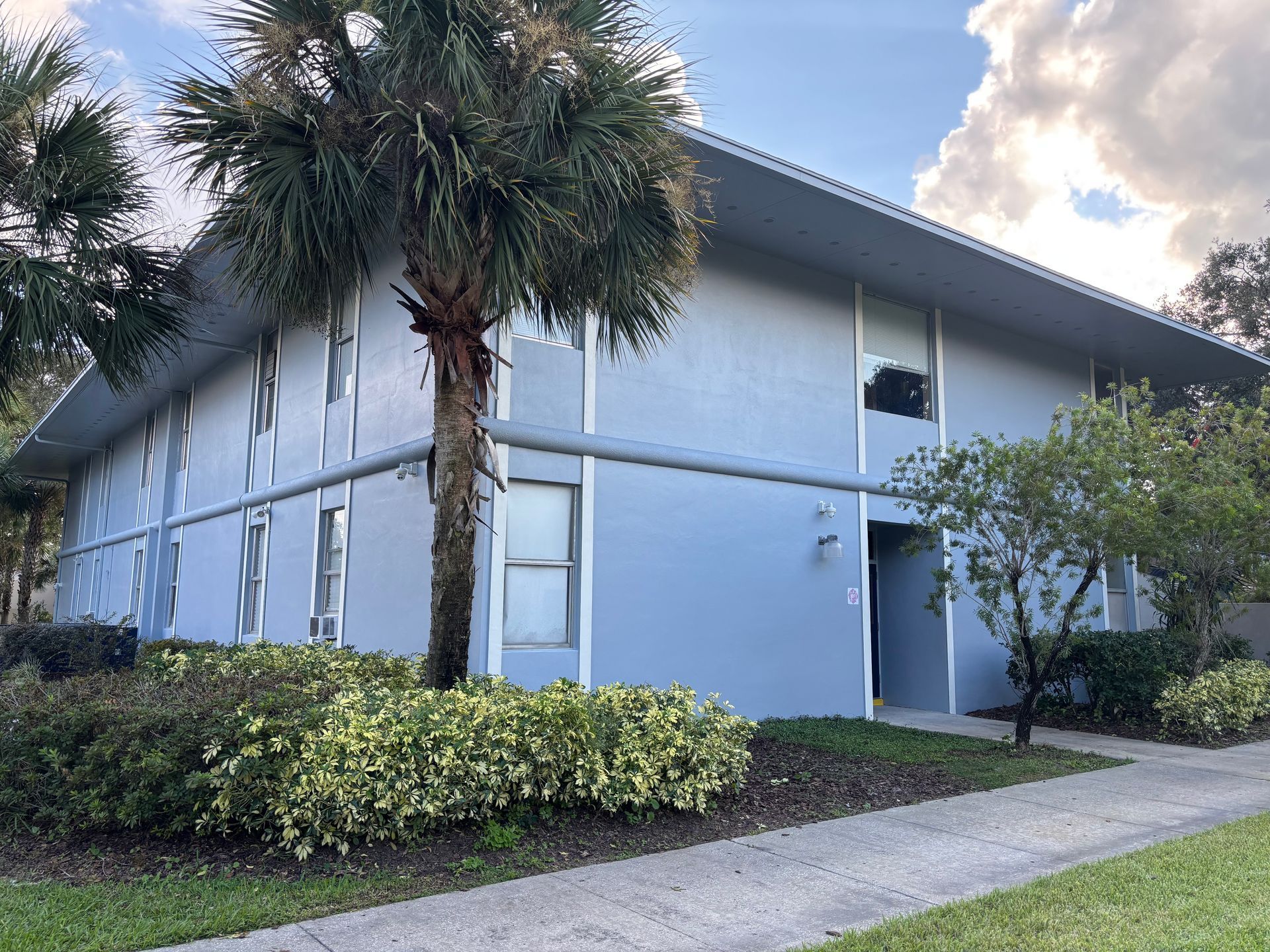 Two-story light blue building with white trim, palm trees, and a sidewalk.