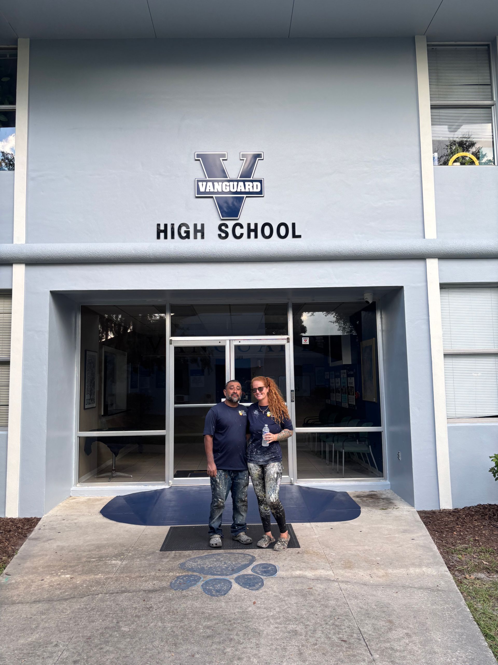 Two people stand in front of Vanguard High School. Gray building, blue lettering, and paw print on ground.