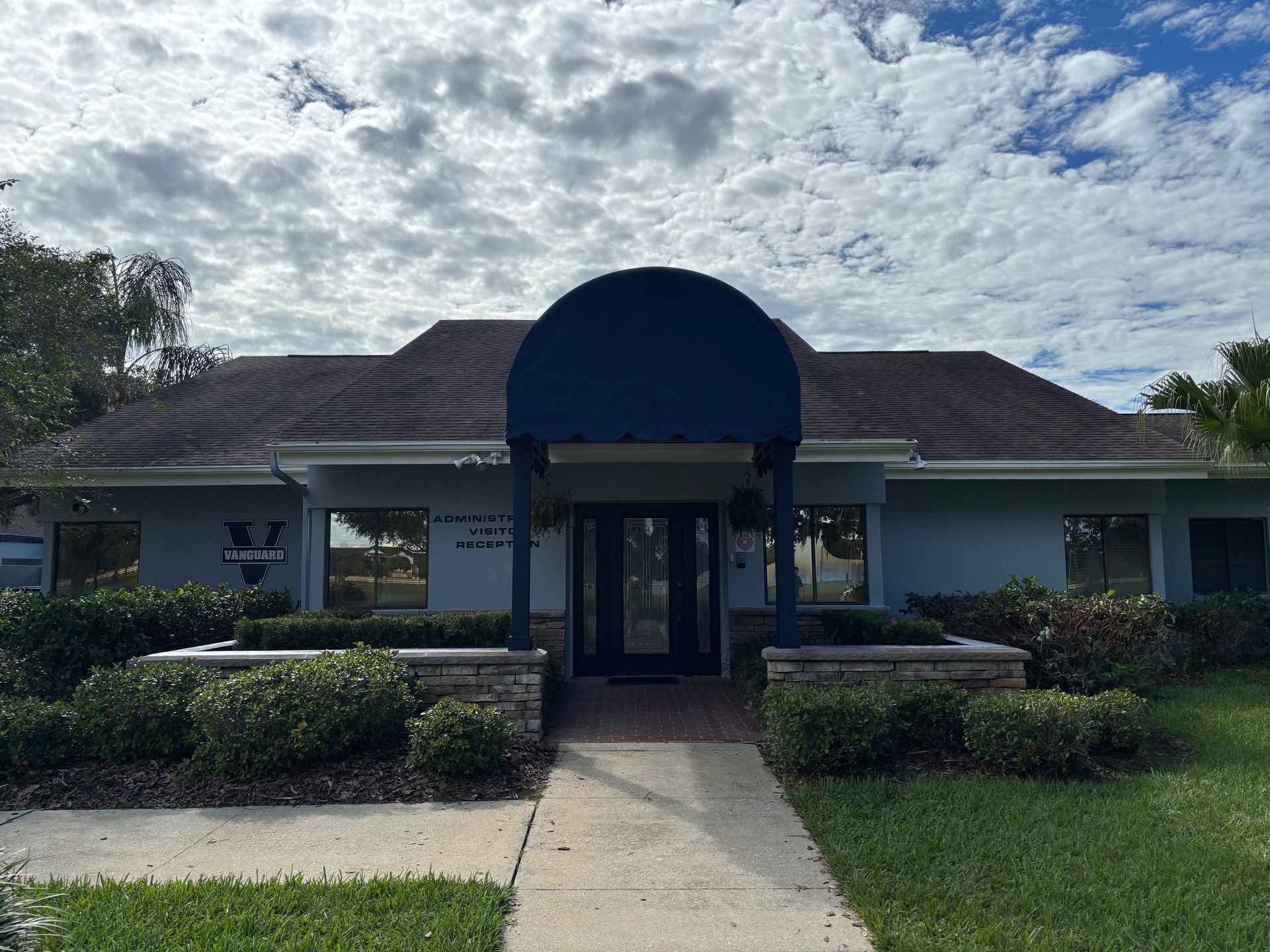 Blue building with dark blue awning over the entrance and a walkway. Green bushes and cloudy sky.