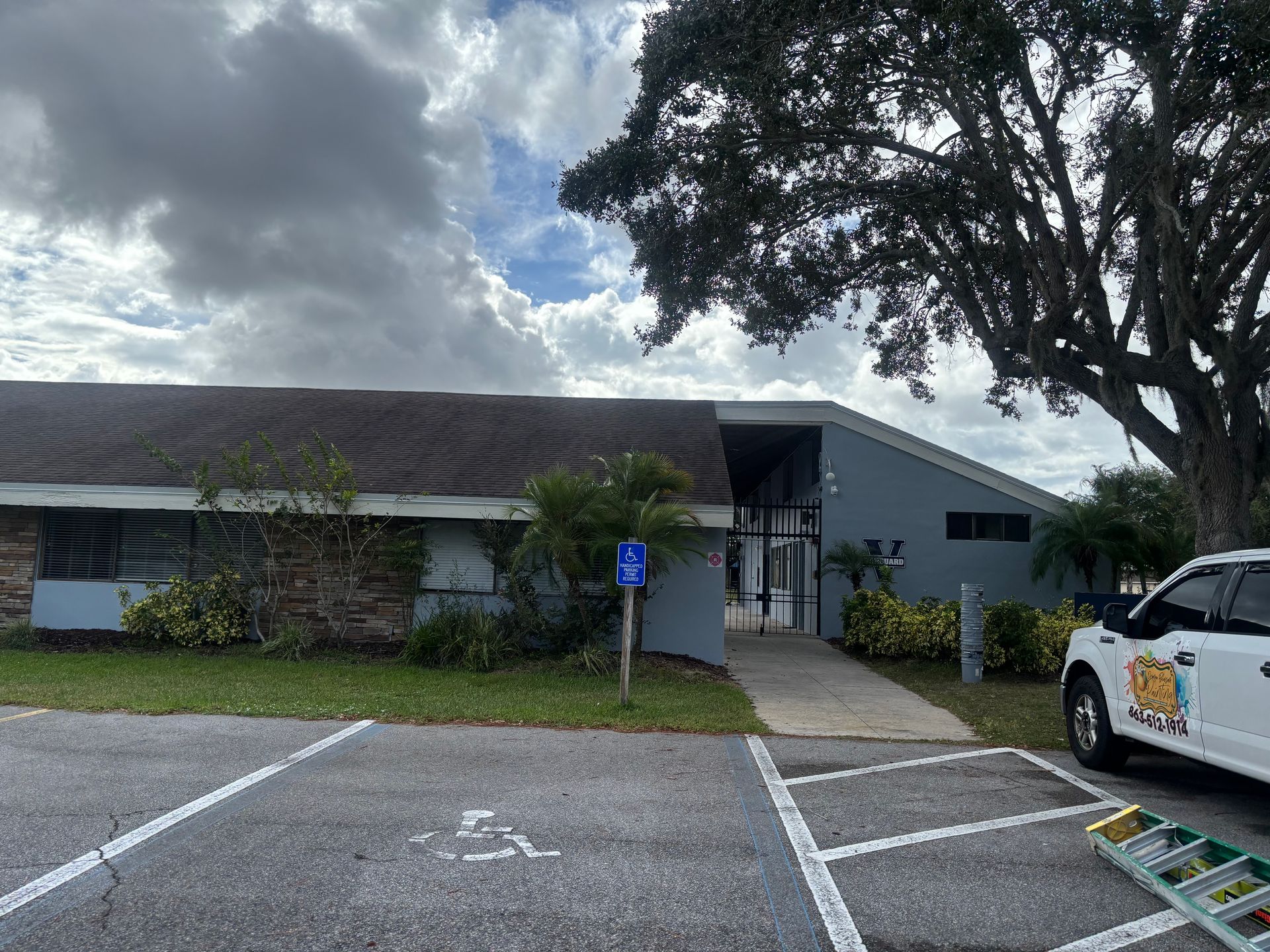 Exterior of a low-rise building with a handicap parking spot. Gray and green facade, tree, and a vehicle.