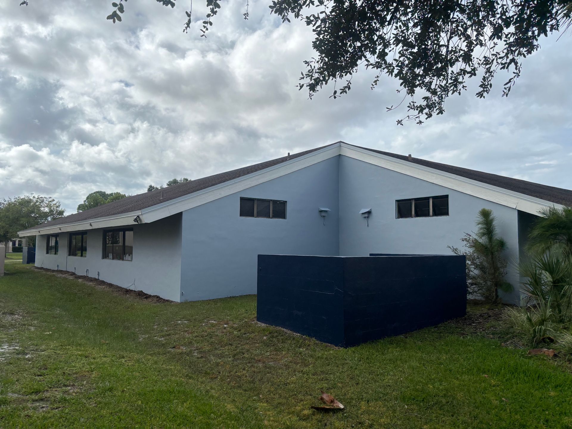 A light blue building with a dark blue structure in front, on a grassy lawn under a cloudy sky.