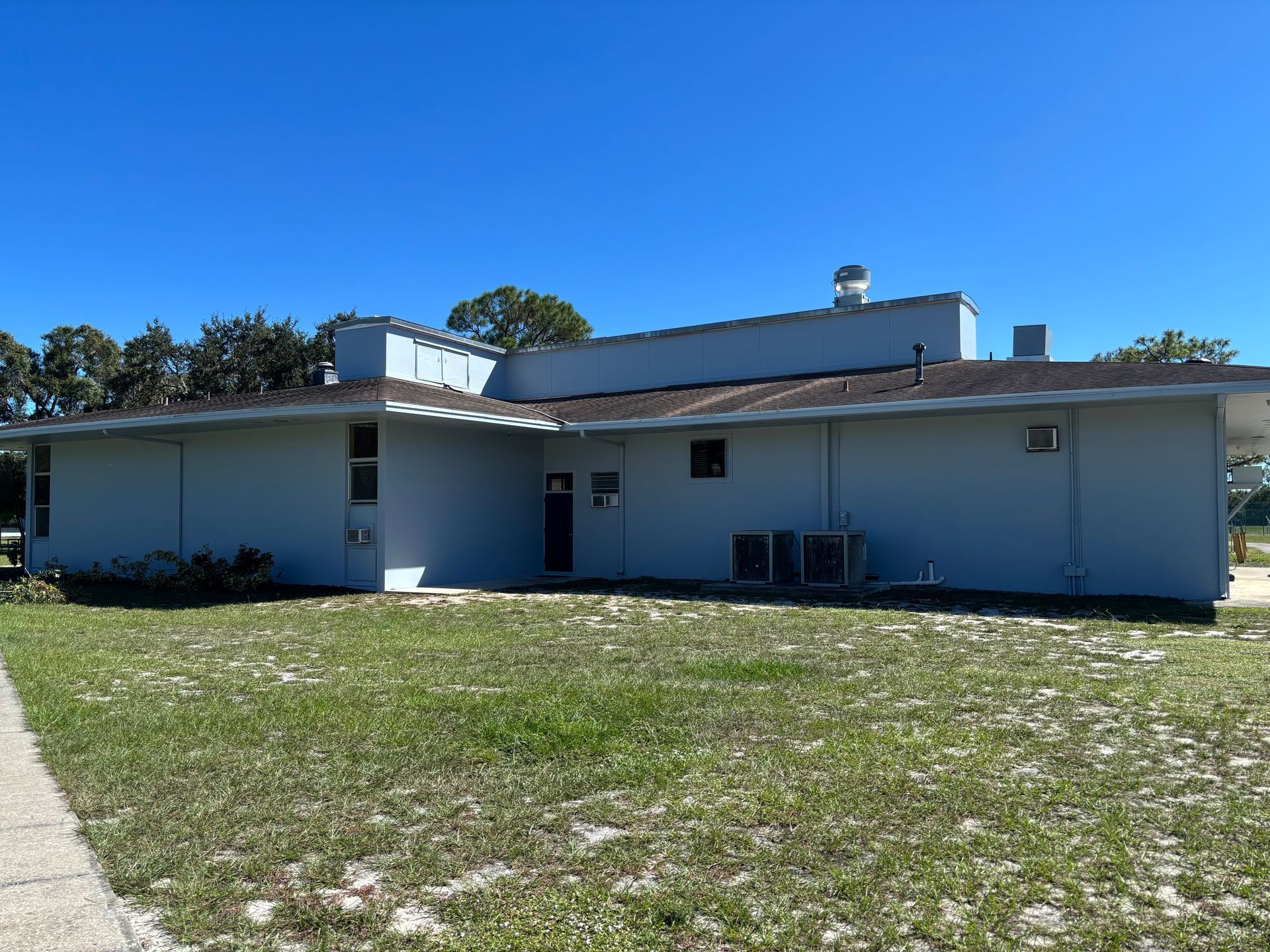 A low, light-blue building with a flat roof and air conditioning units in a grassy area under a clear blue sky.