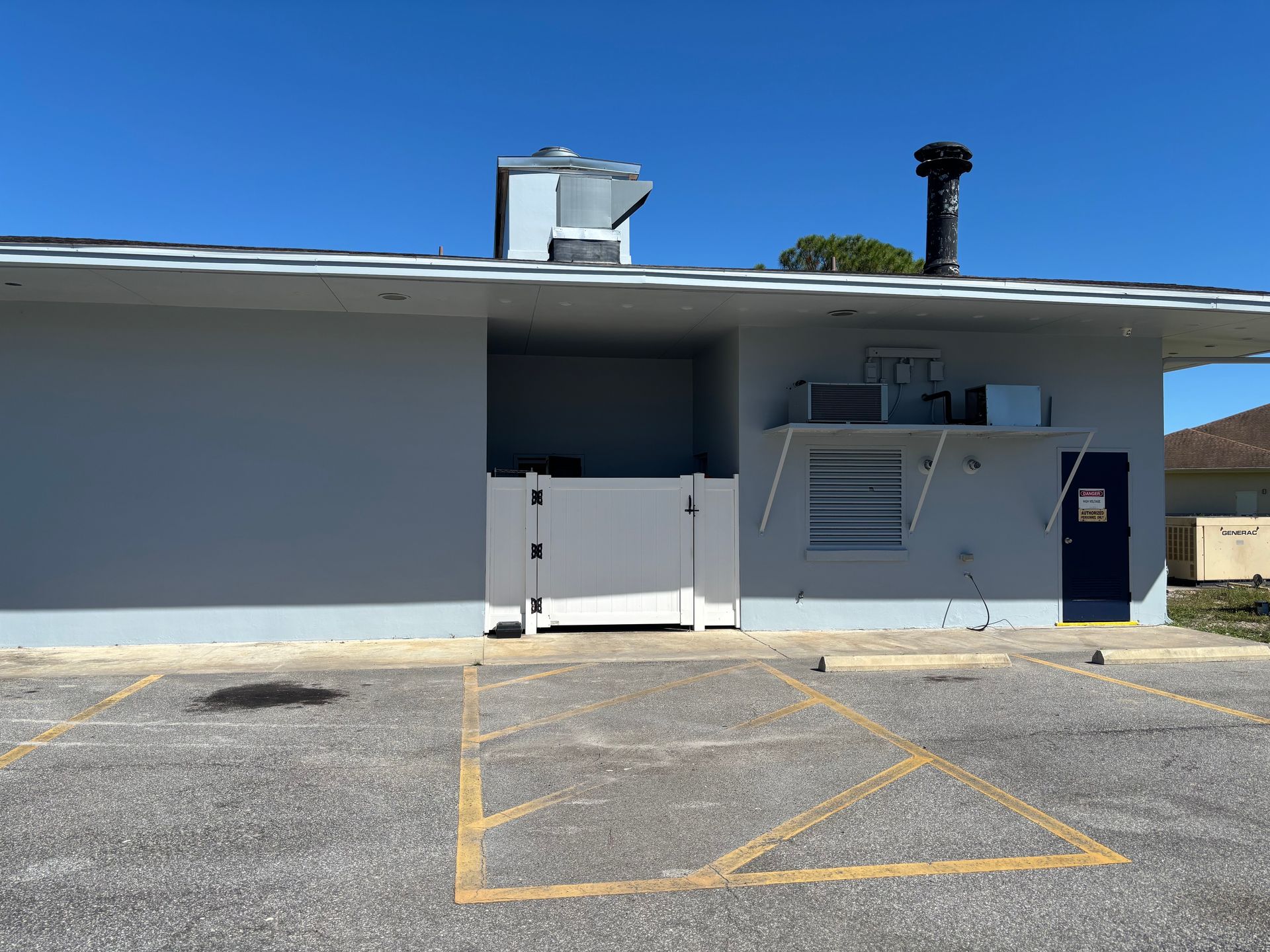 Gray building with white gate, vent, and blue door, in a parking lot on a sunny day.