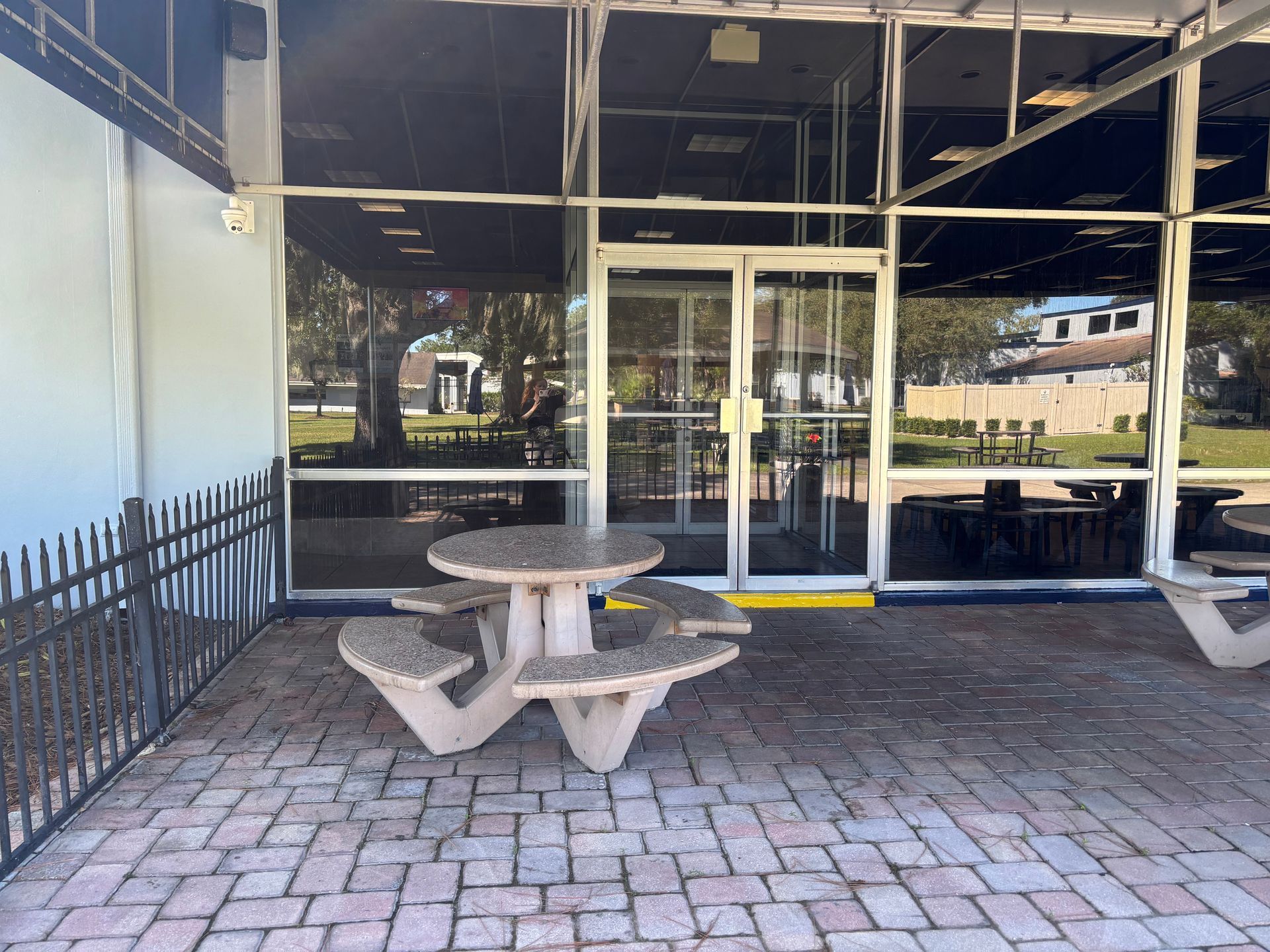 Picnic table in front of a building with glass doors and windows, brick patio, black fence.