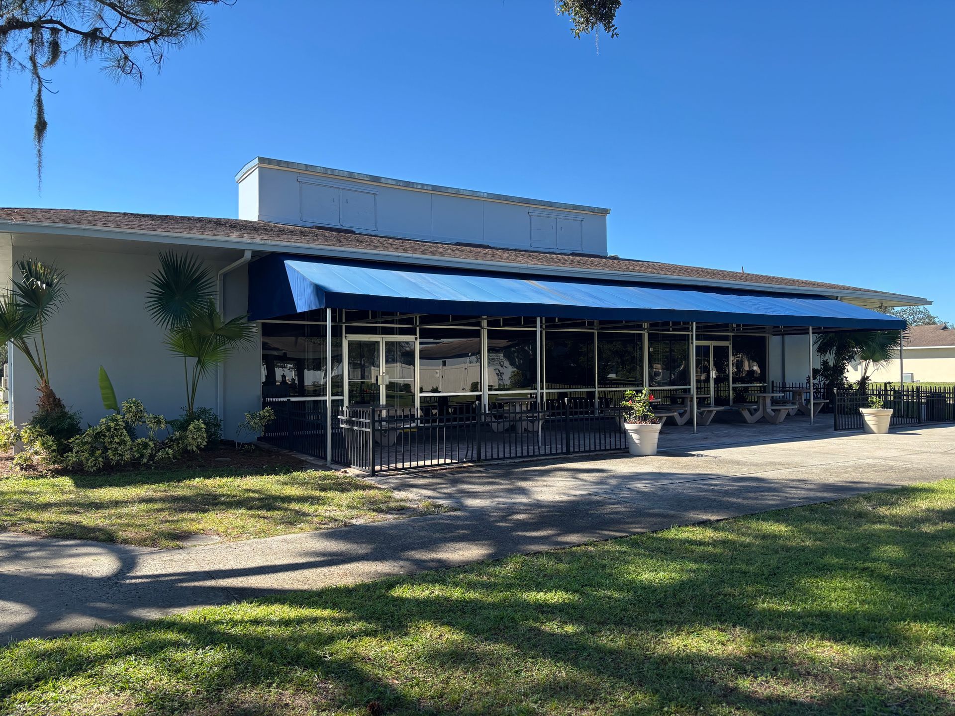 Exterior of a building with a blue awning and glass windows. Tables and chairs are visible outside.