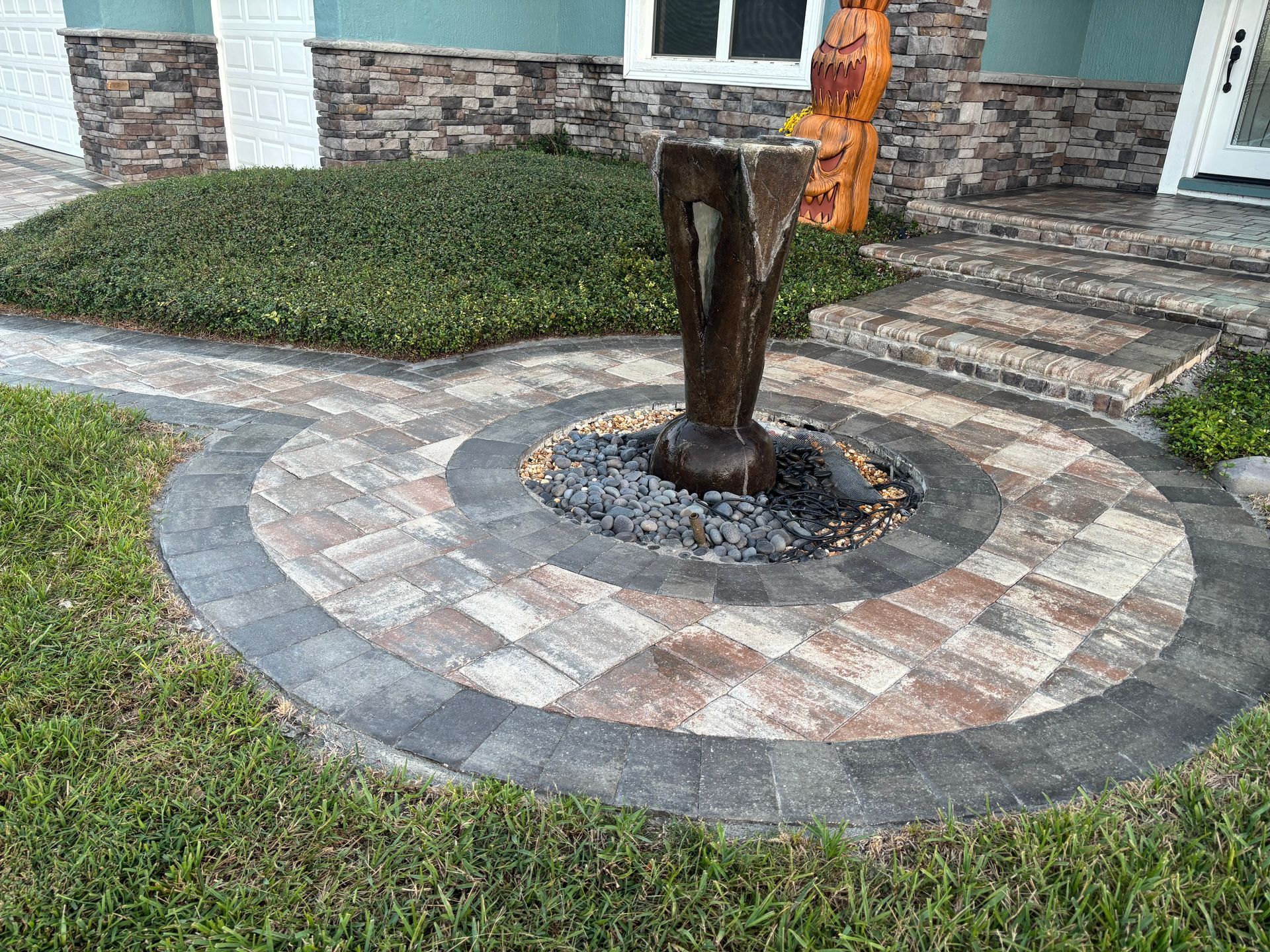 Stone fountain in a brick and stone paved circle, surrounded by grass and shrubs, near a house.
