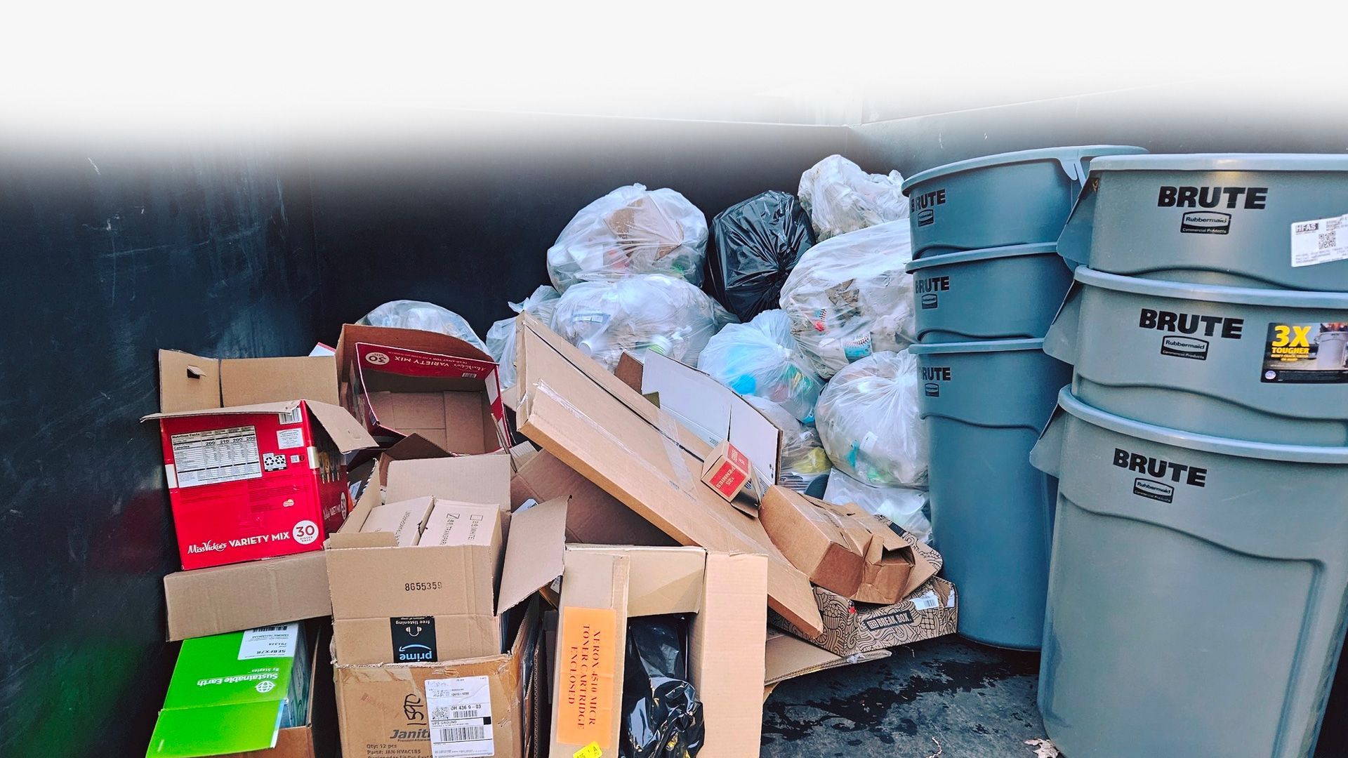 Dumpster overflowing with cardboard boxes, trash bags, and stacked gray bins.