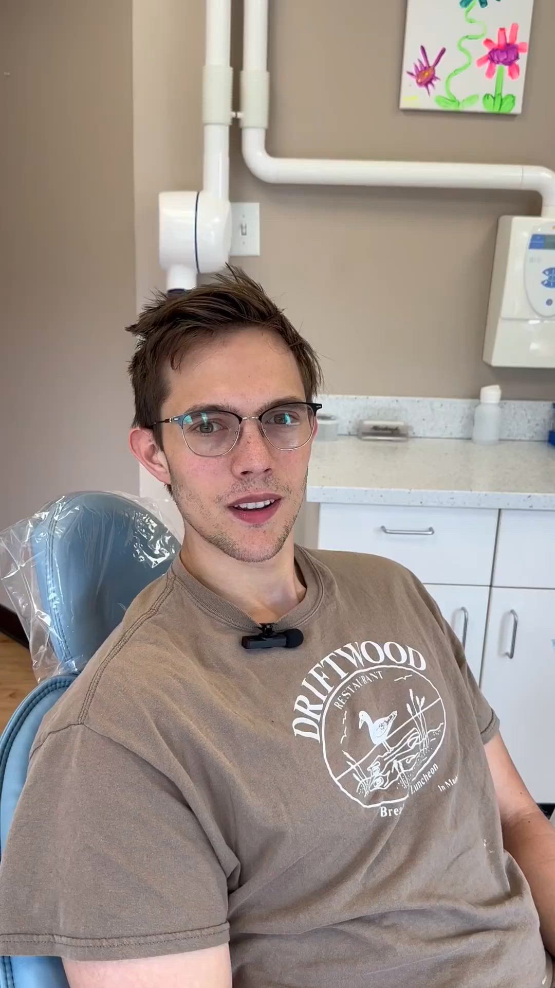 A man is sitting in a dental chair wearing glasses and a brown shirt.