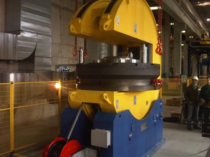 Un grande macchinario industriale, giallo e blu, viene sollevato. Gli operai osservano in un ambiente industriale.